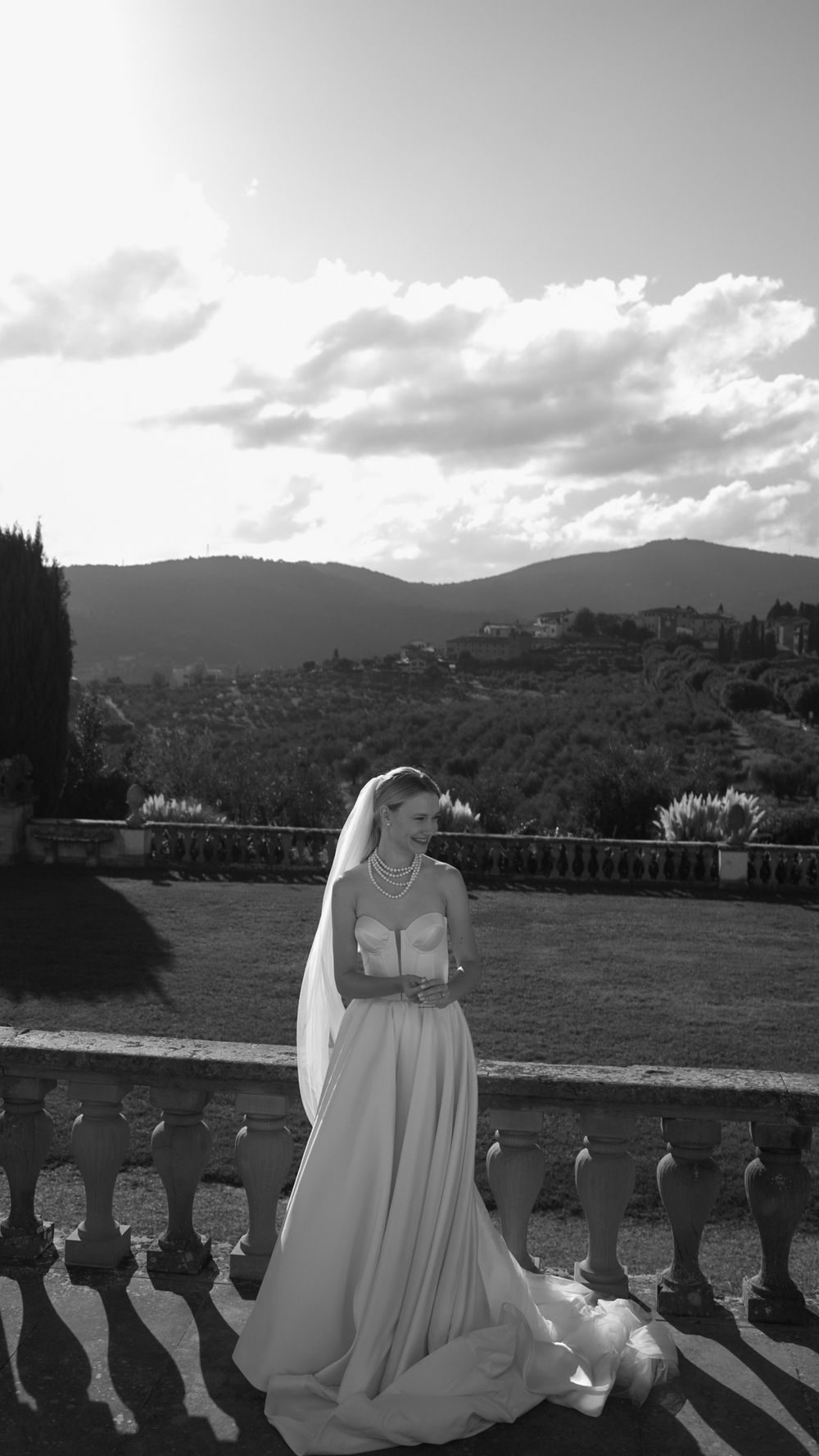 Black and white photo of a bride in a strapless gown and veil standing on a balustrade terrace.