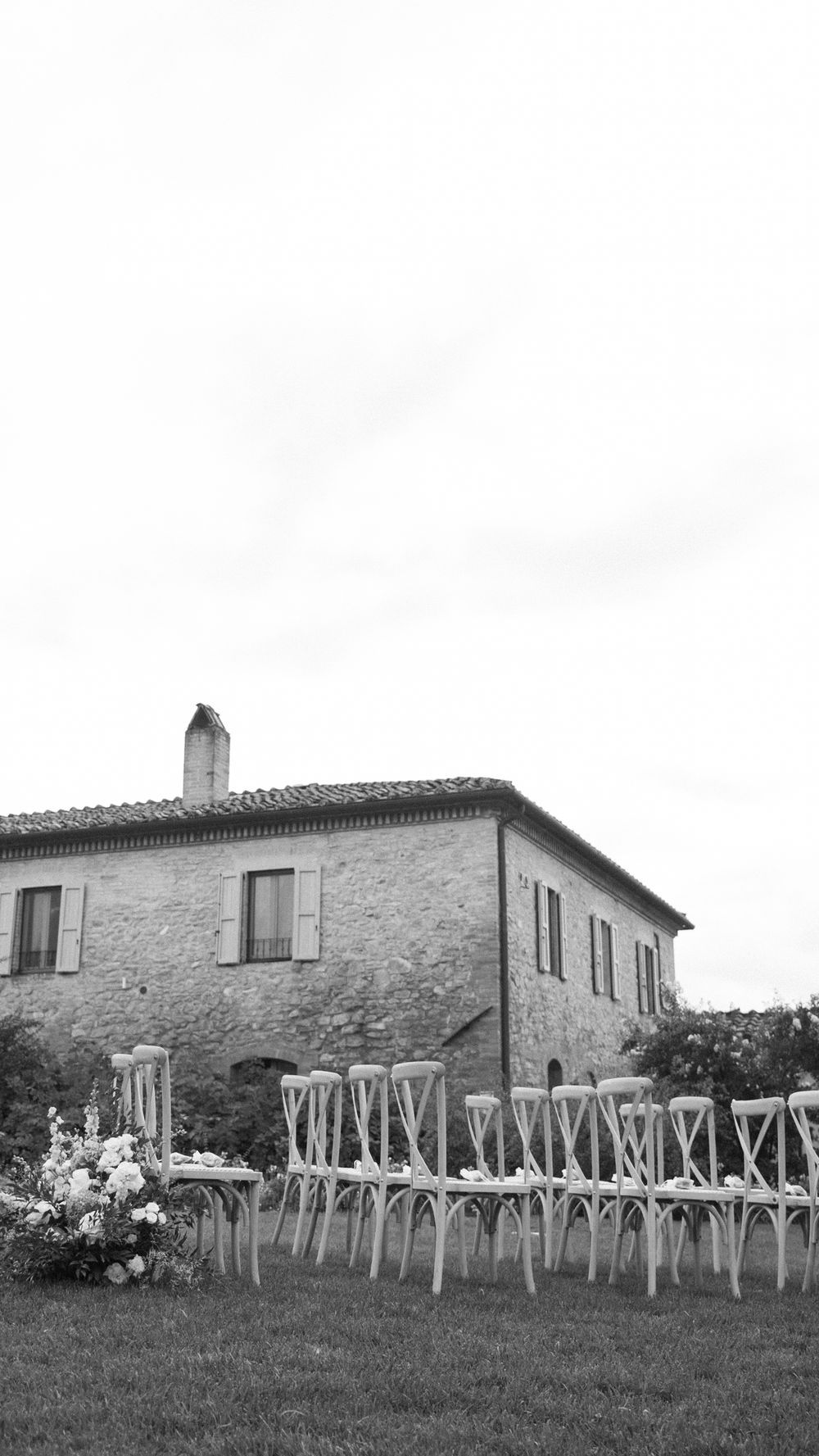 Black and white photo of rustic stone villa with rows of wooden chairs set for outdoor wedding ceremony.