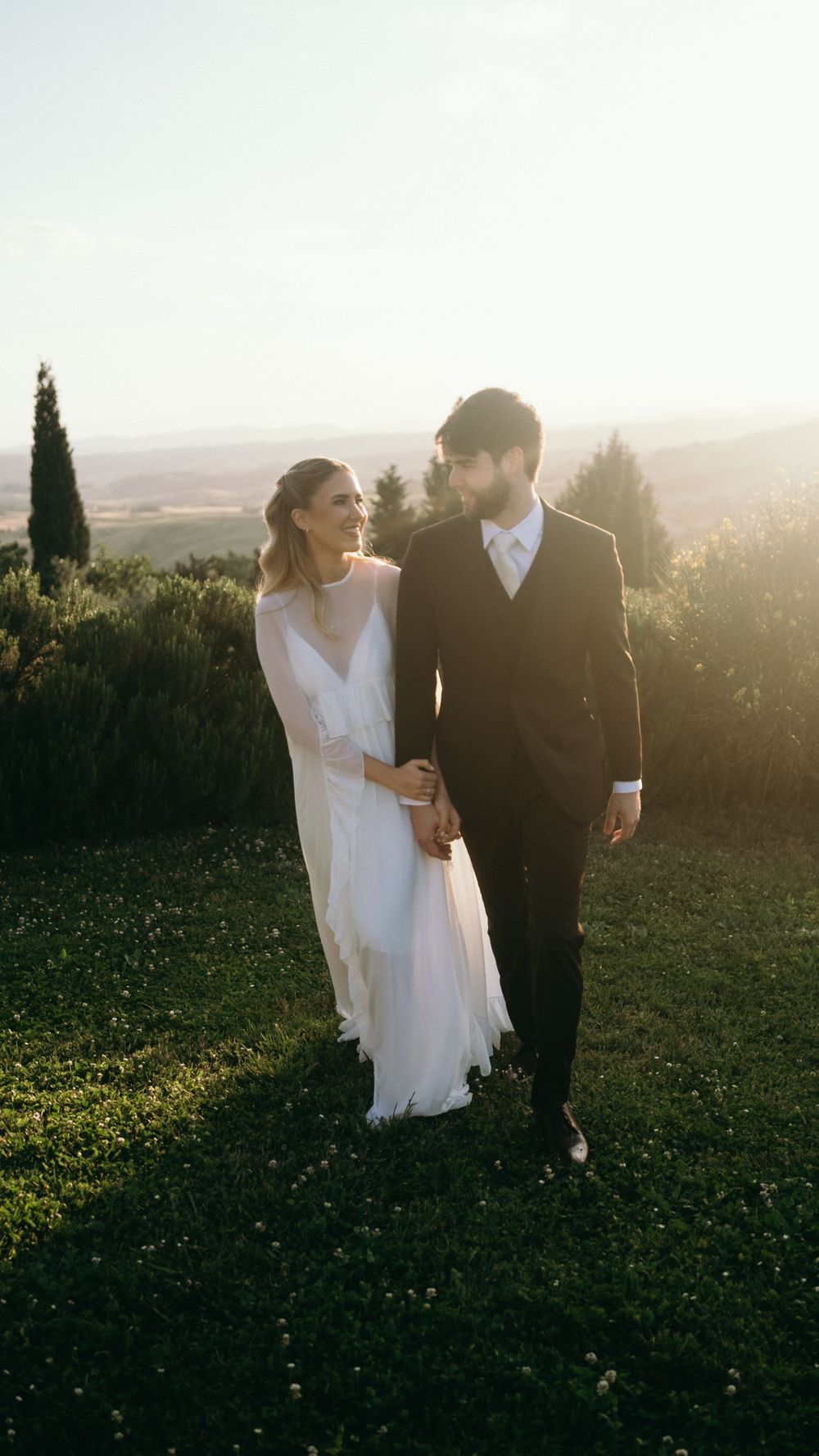 Bride and groom walking hand in hand through lush green gardens at golden hour during outdoor wedding.