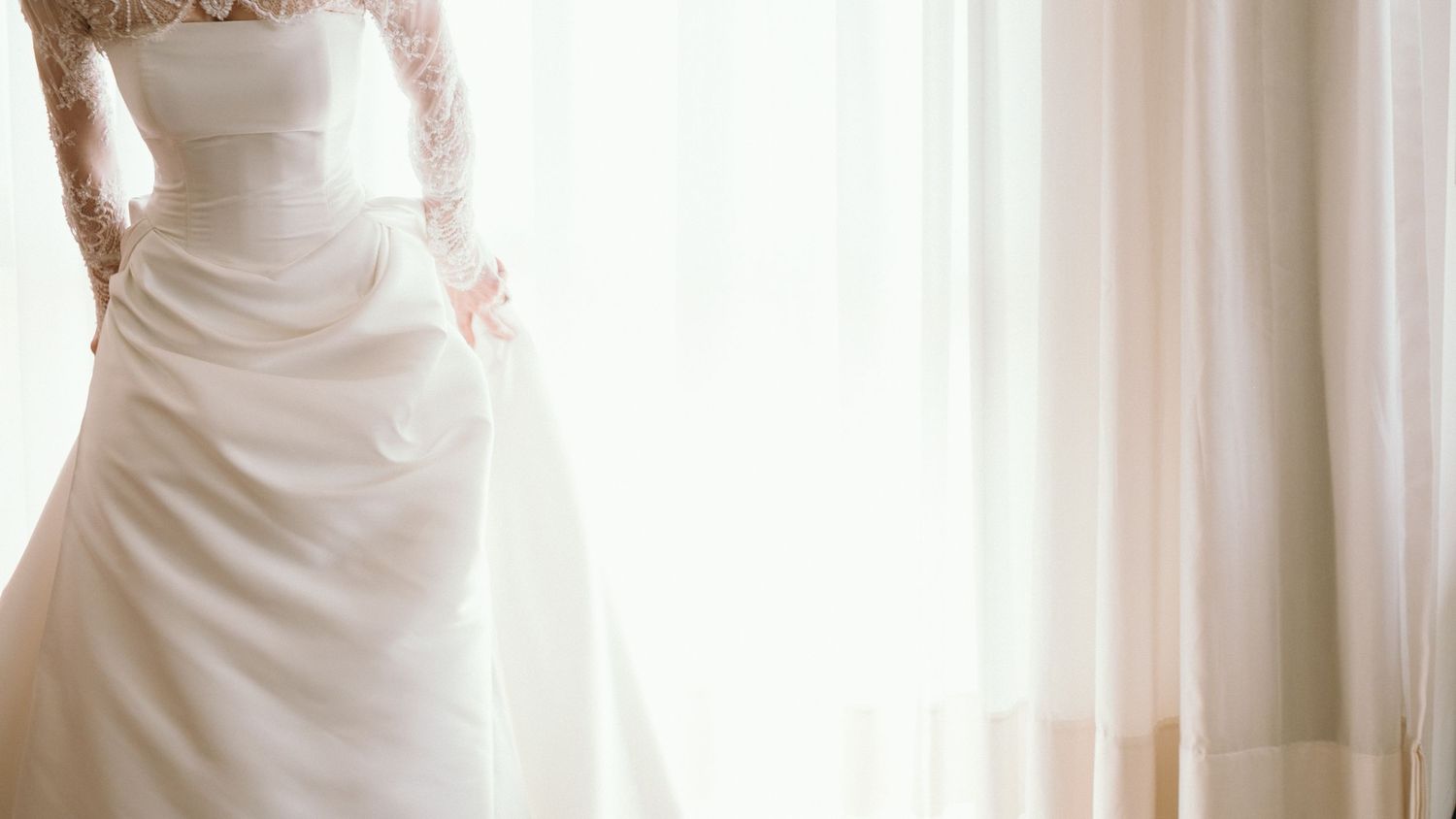 A bride in a strapless white satin wedding gown with lace sleeves stands by sheer curtains.