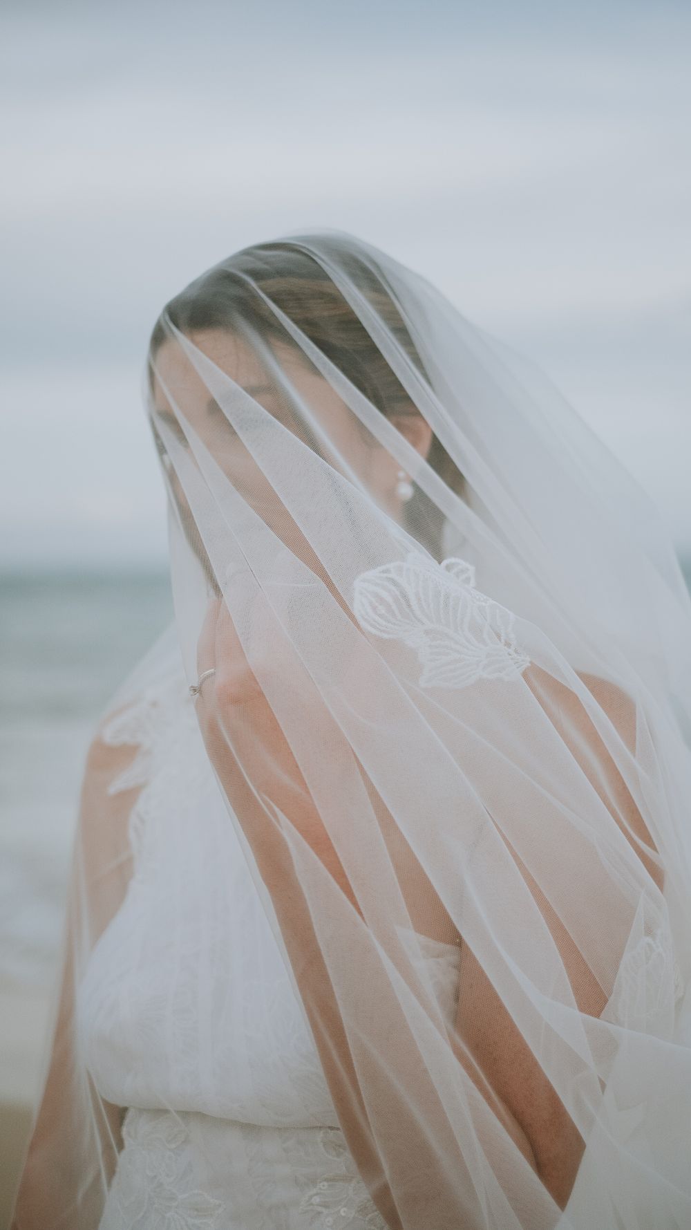 Bride in white wedding dress with embroidered veil blowing across her face on a beach.