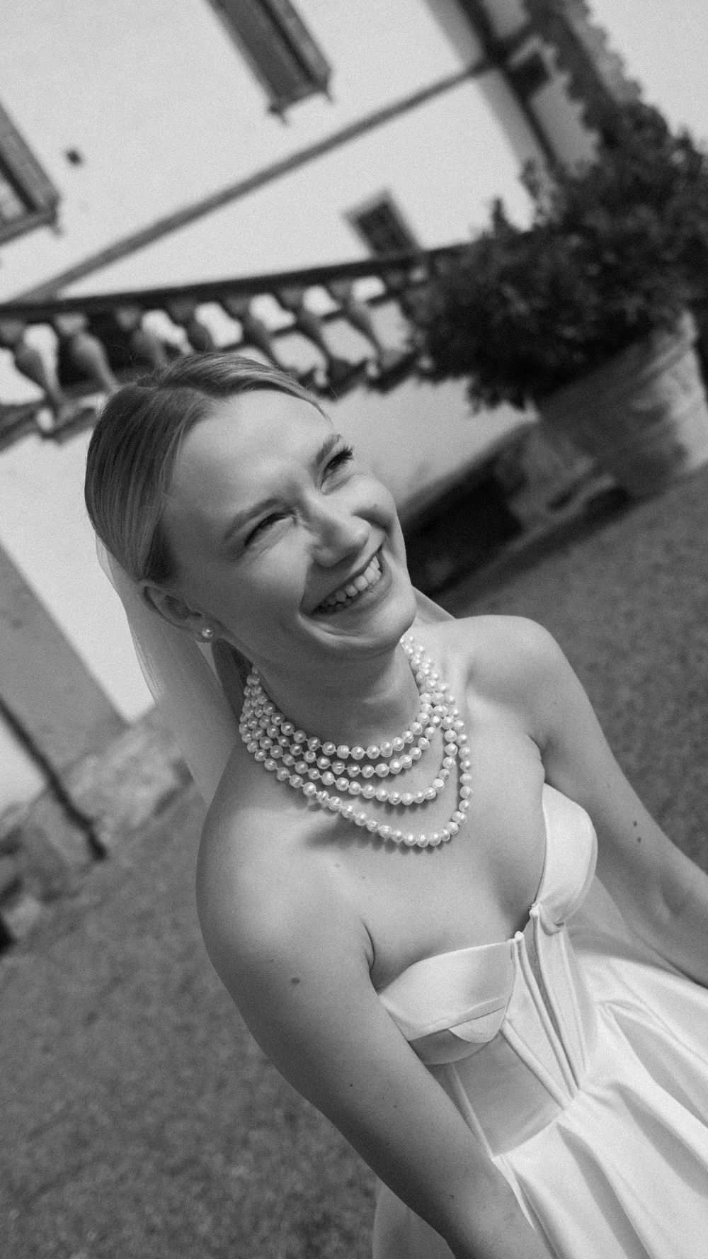 Smiling bride wearing layered pearl necklace in strapless white wedding gown, black and white photo.