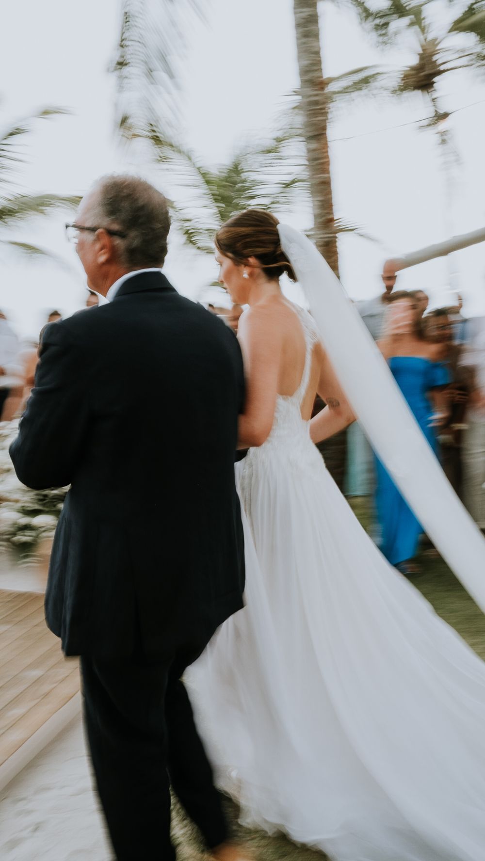 Motion-blurred photo of bride in white gown walking with father during outdoor ceremony.