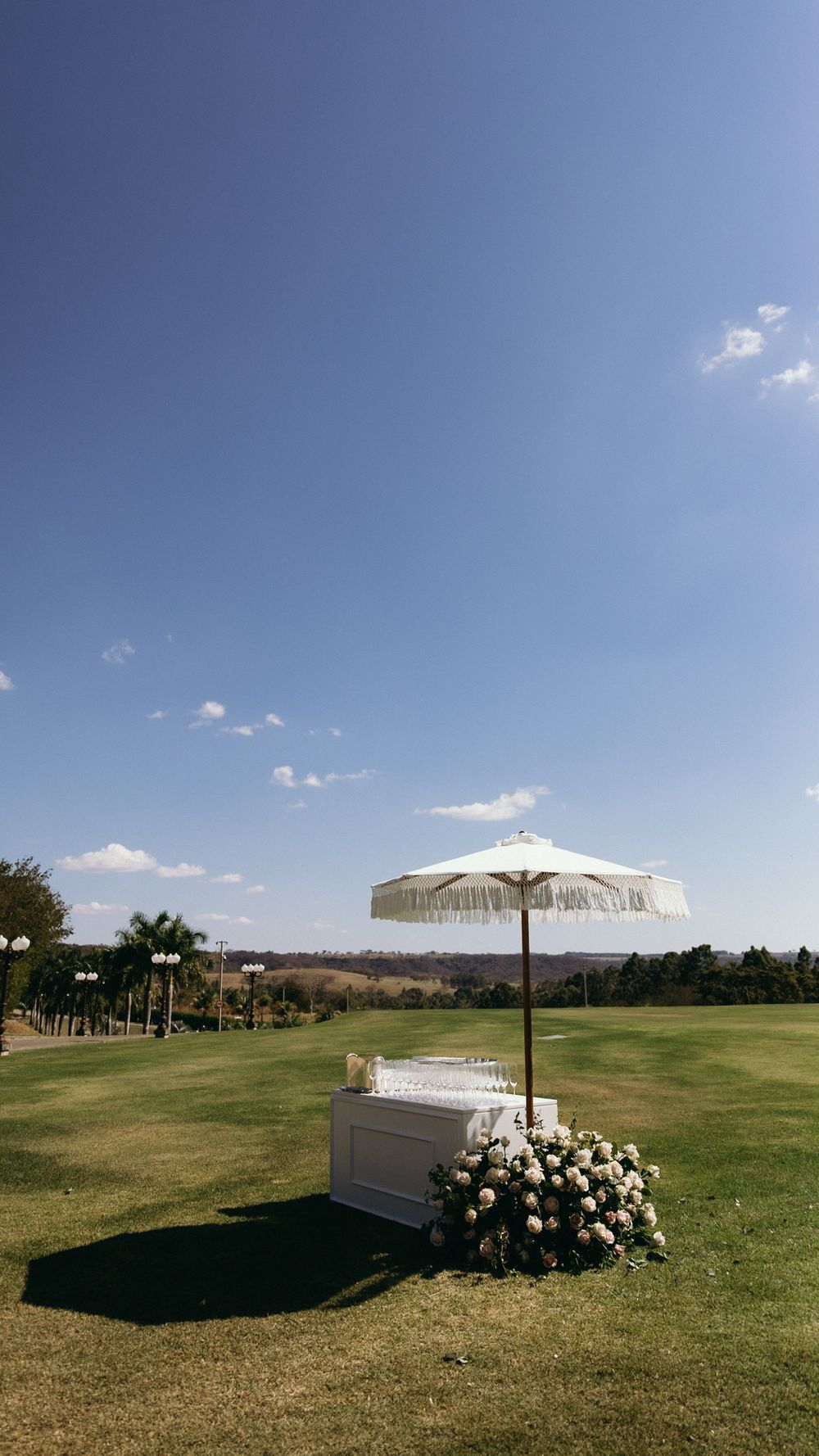 White fringed umbrella over a floral-decorated bar station on a sunny green lawn under blue sky.
