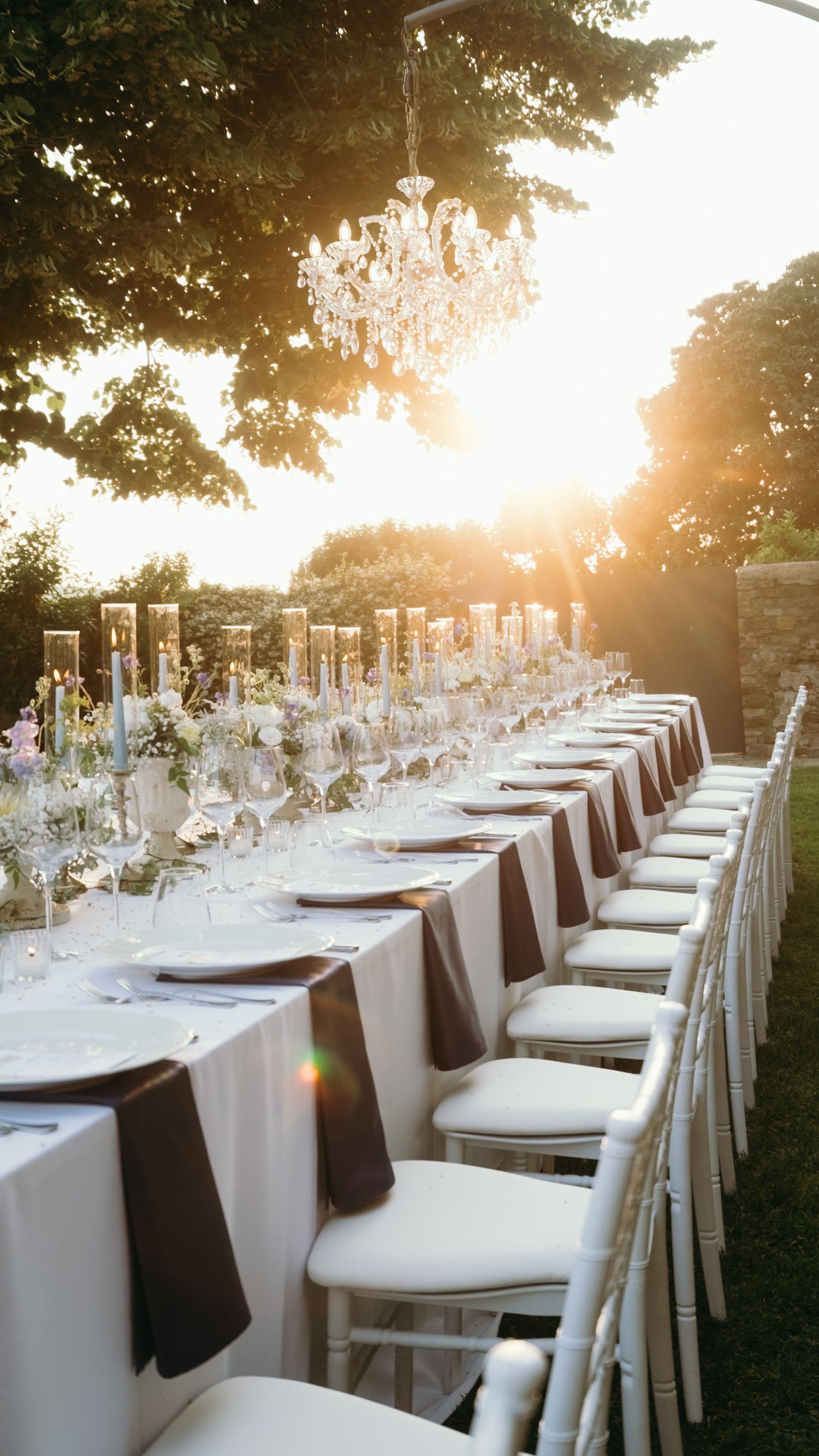 Elegant outdoor wedding reception table set at golden hour with crystal chandelier and white chiavari chairs.