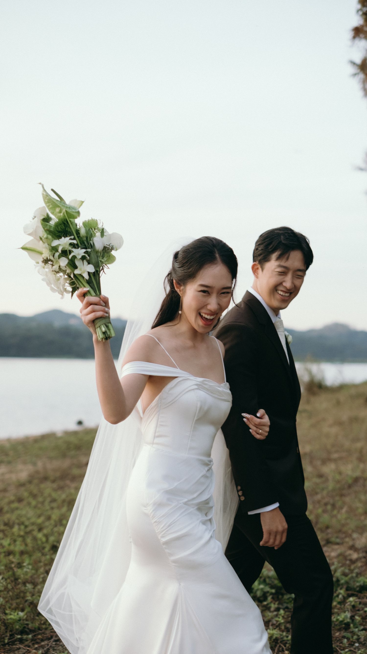 Joyful Asian bride in off-shoulder gown raising bouquet while walking with groom by lake.