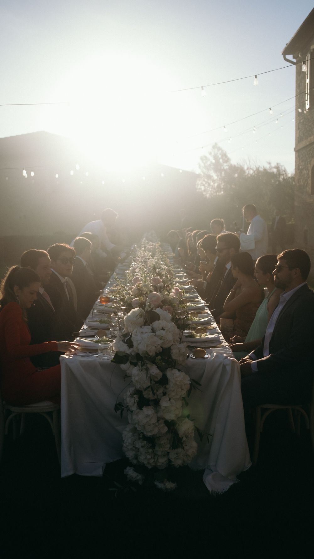 Elegant outdoor wedding reception with long decorated table, floral centerpieces, and guests dining at golden hour.