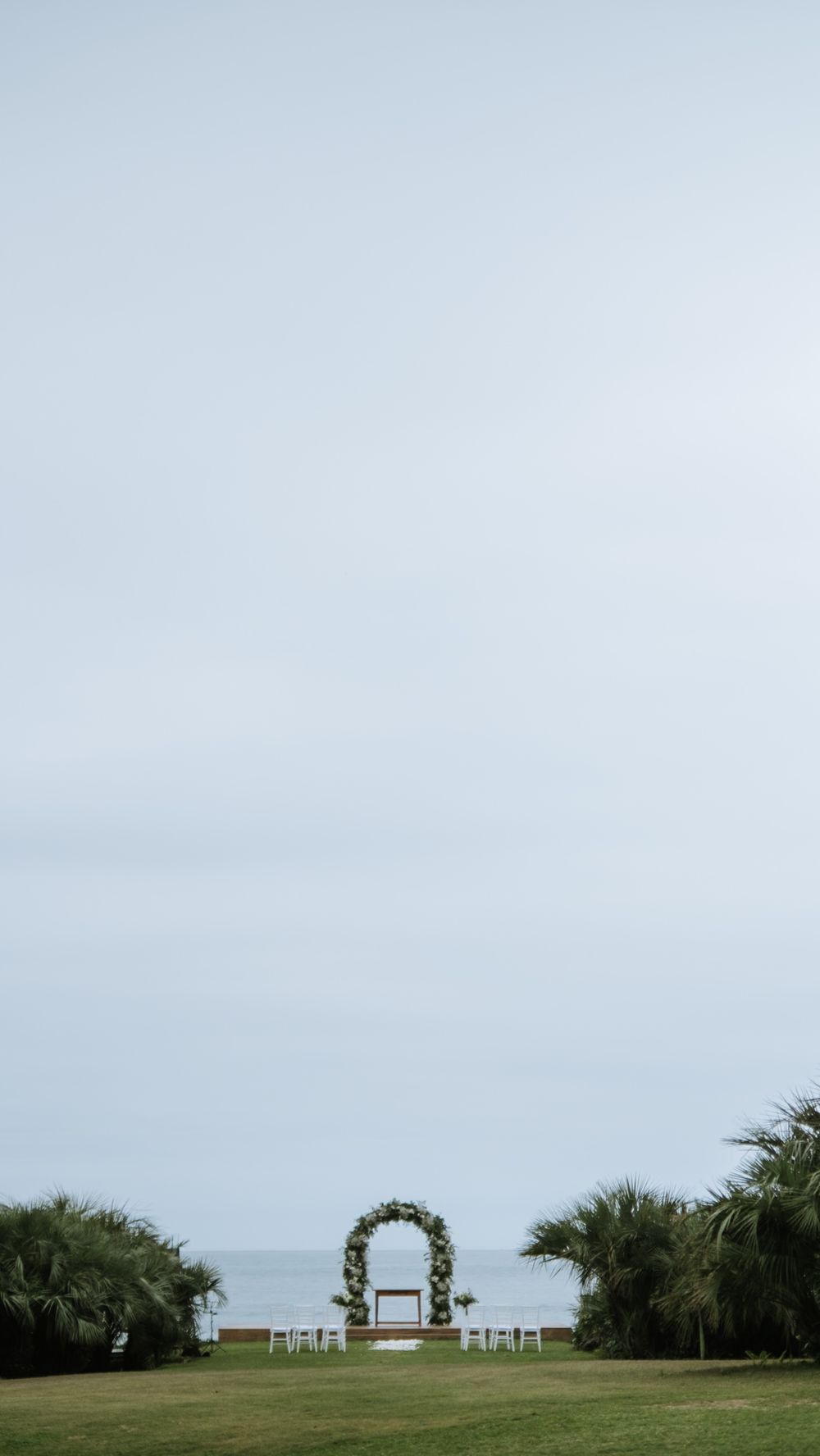 Minimalist outdoor wedding ceremony arch with floral decor overlooking ocean and palm trees.
