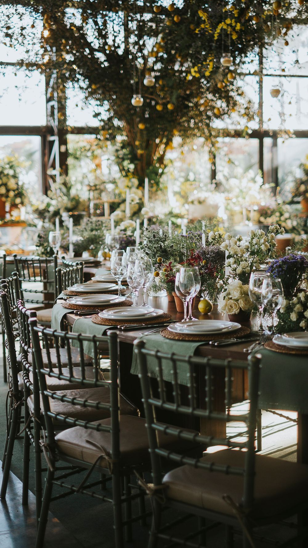 Elegant wedding reception table with sage green runner, candles, wildflower arrangements under lush floral ceiling.