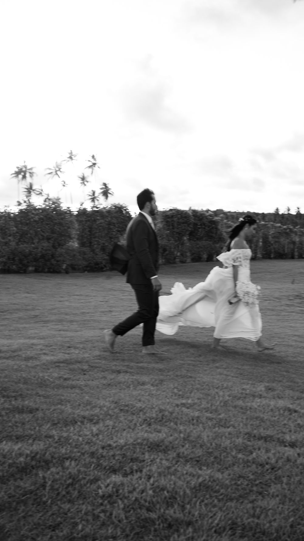 Black and white photo of bride and groom running barefoot across a grassy field with palm trees.