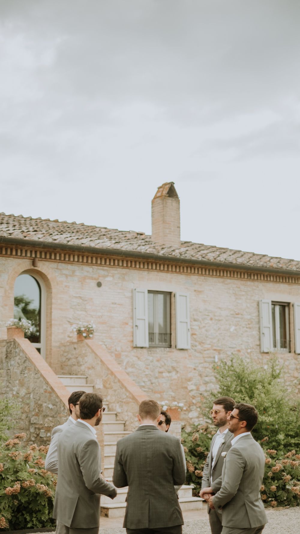 Groomsmen in grey suits gather outside rustic Tuscan stone villa on wedding day.