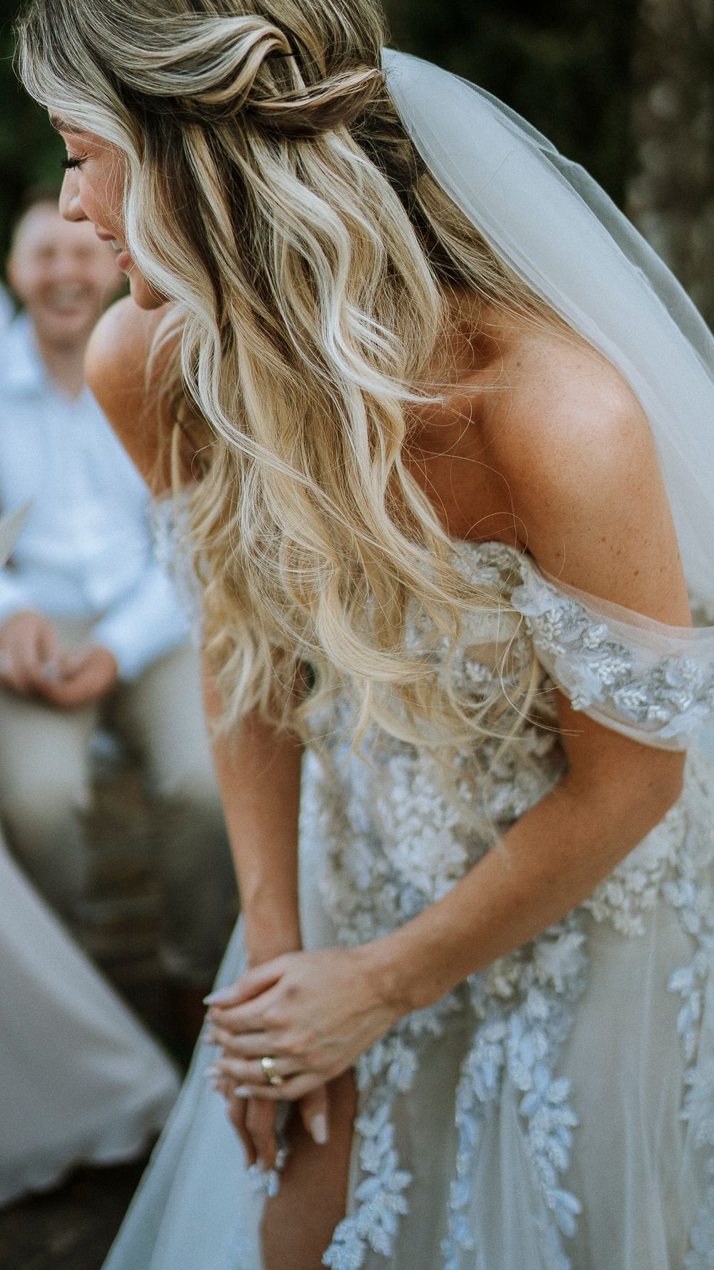 Bride with long wavy blonde hair in floral lace off-shoulder wedding gown during outdoor ceremony.