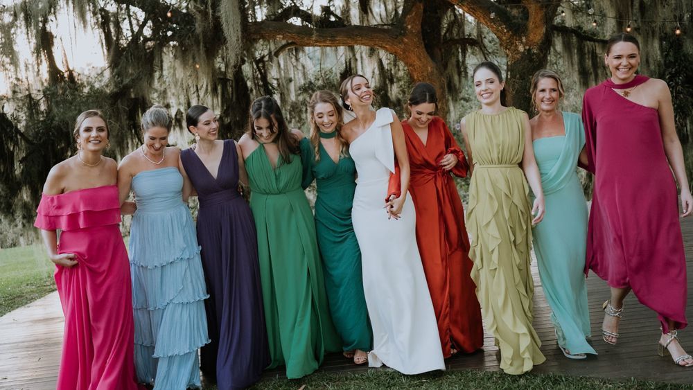 A bride in white stands surrounded by bridesmaids in colorful rainbow gowns under Spanish moss-draped trees.