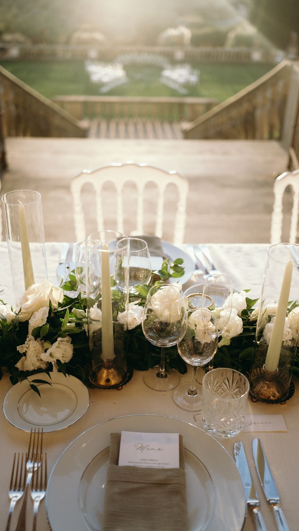 Elegant wedding table setting with white roses, candles, crystal glasses, and greenery garland.