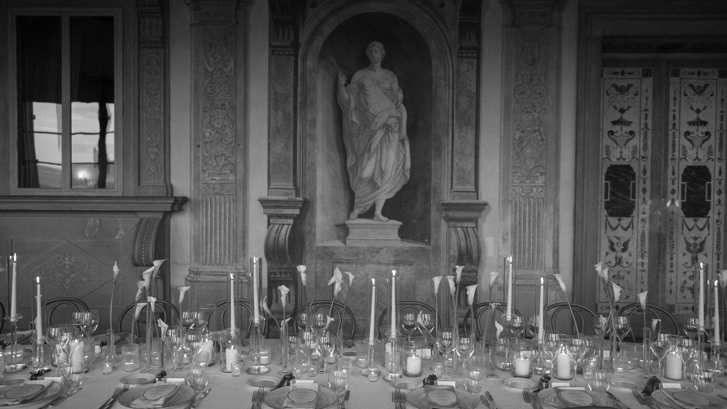 Elegant black and white photo of a formal dining table set with candles beneath a classical statue in an ornate hall.