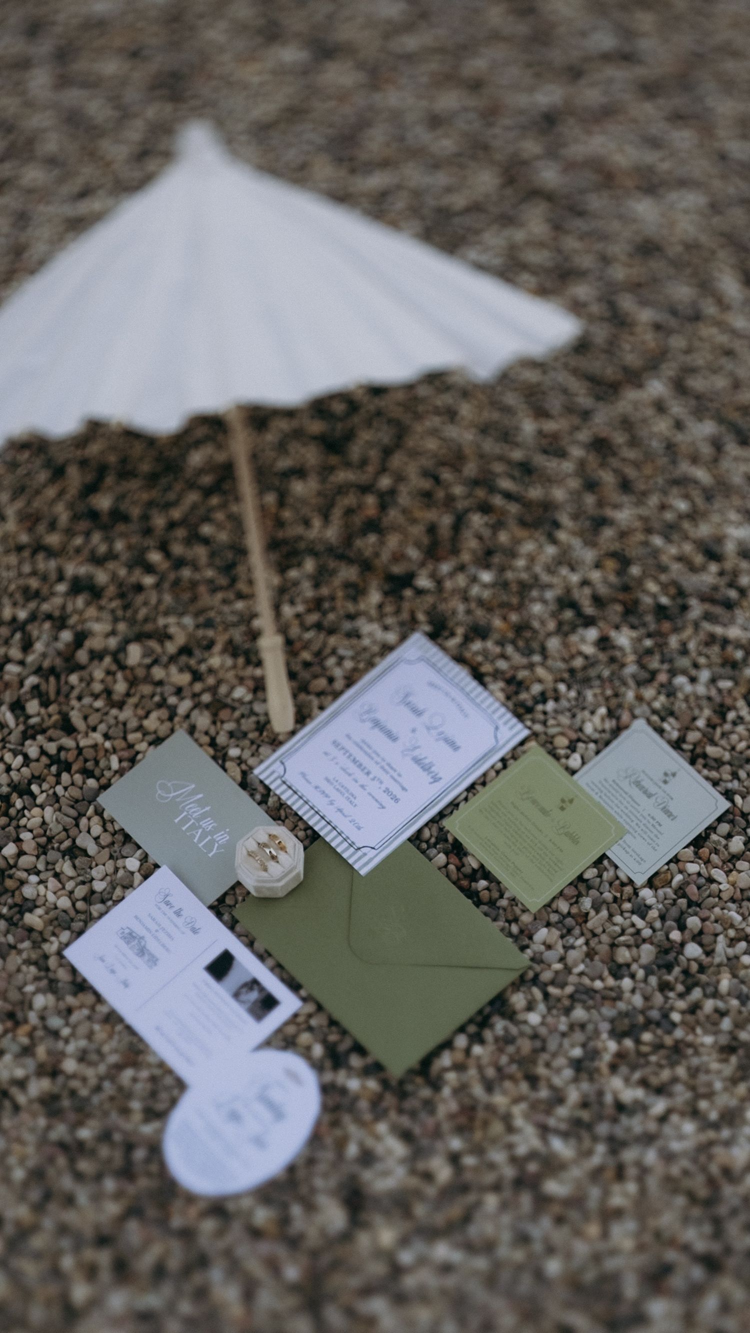 Wedding stationery suite with green envelopes and white parasol arranged on gravel ground.