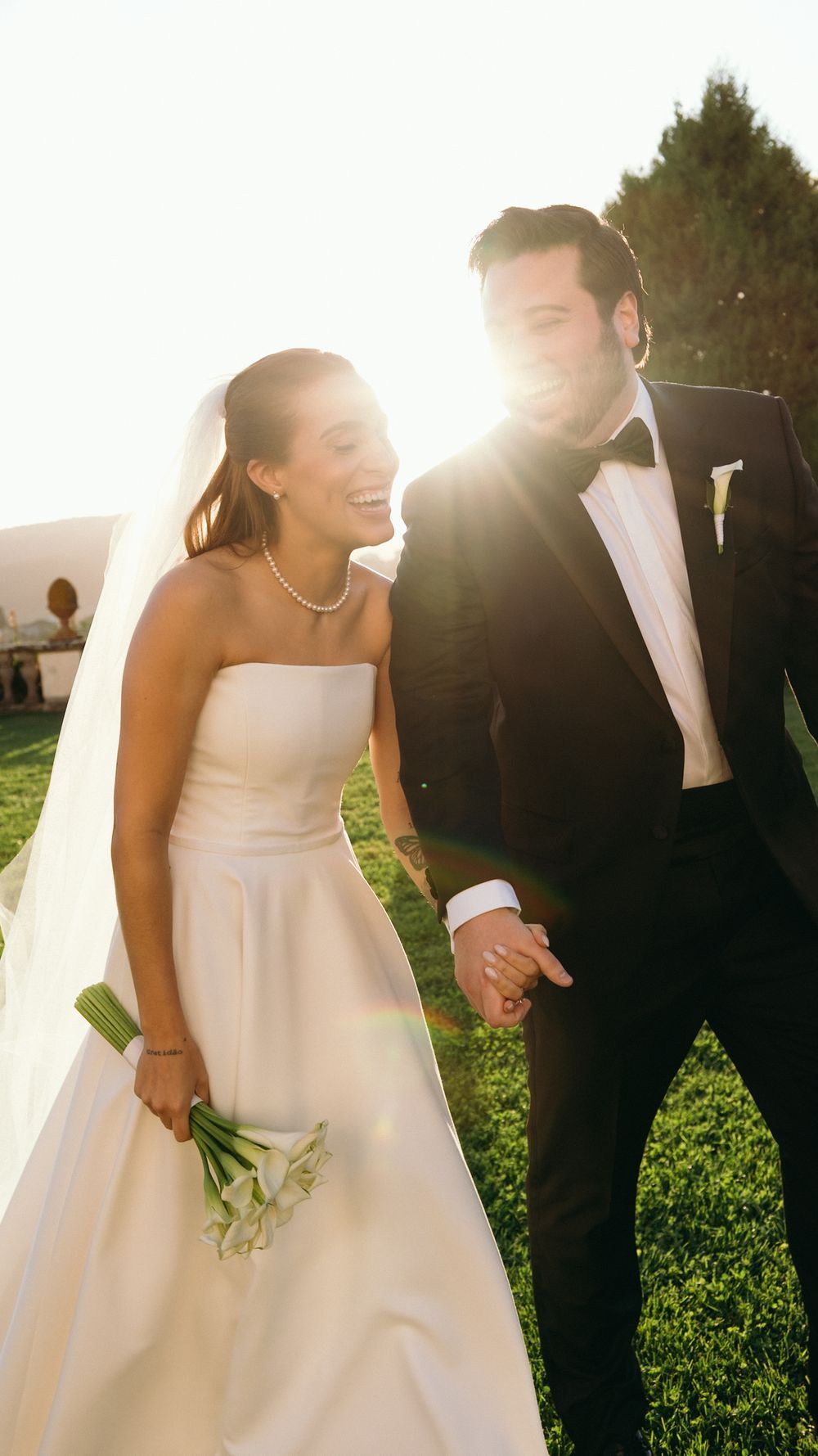 Smiling bride in strapless white gown and groom in black tuxedo walk hand in hand in golden sunlight.
