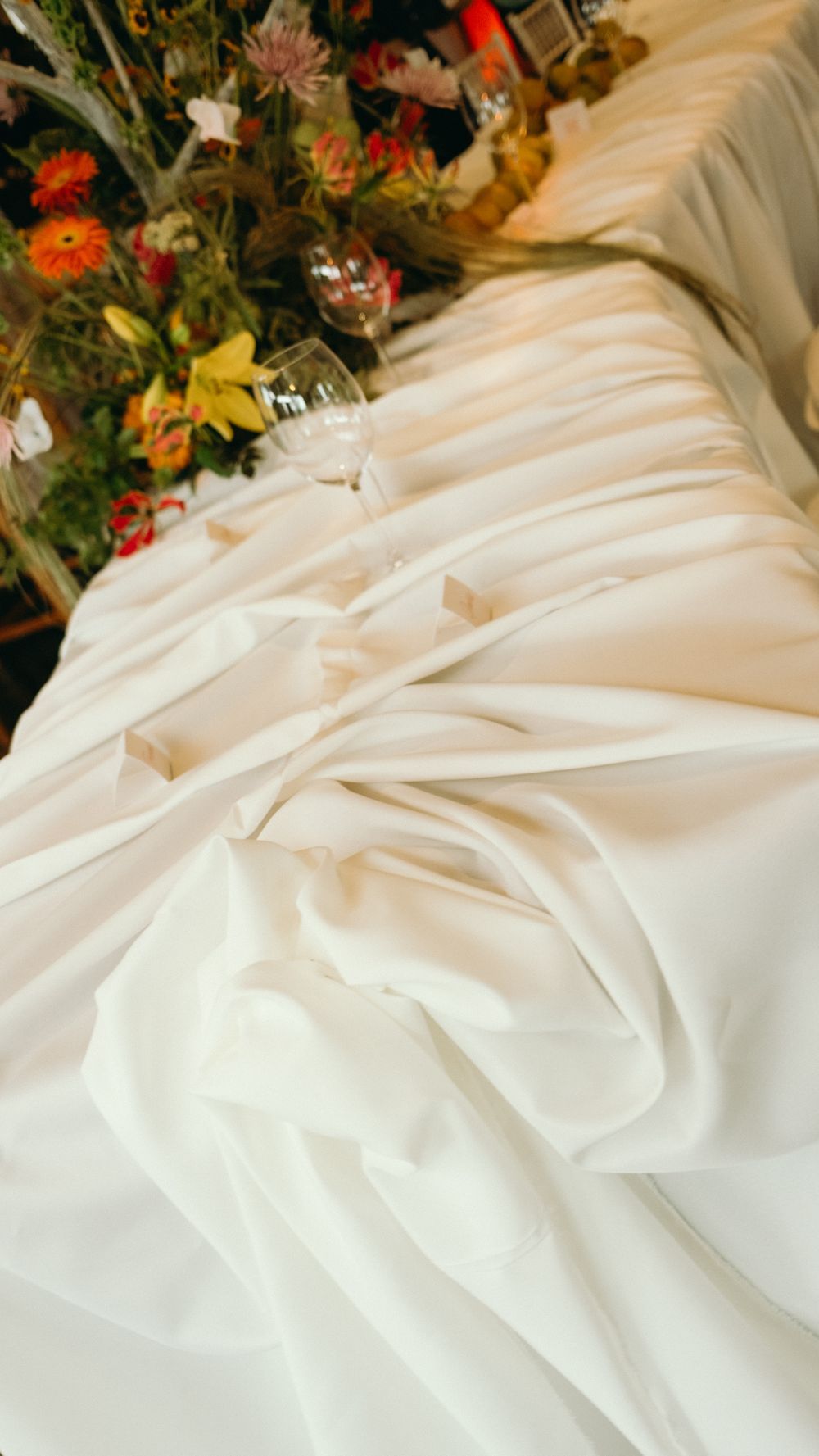 Close-up of elegant white satin wedding dress fabric with floral centerpiece in background.