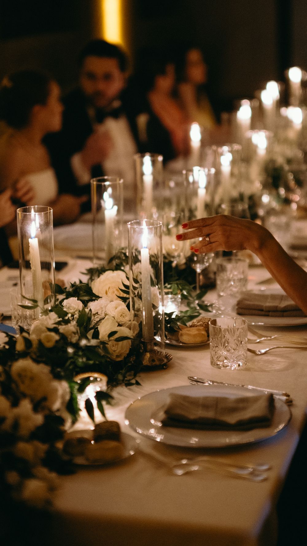 Candlelit wedding reception table with white floral arrangements and guests in background.