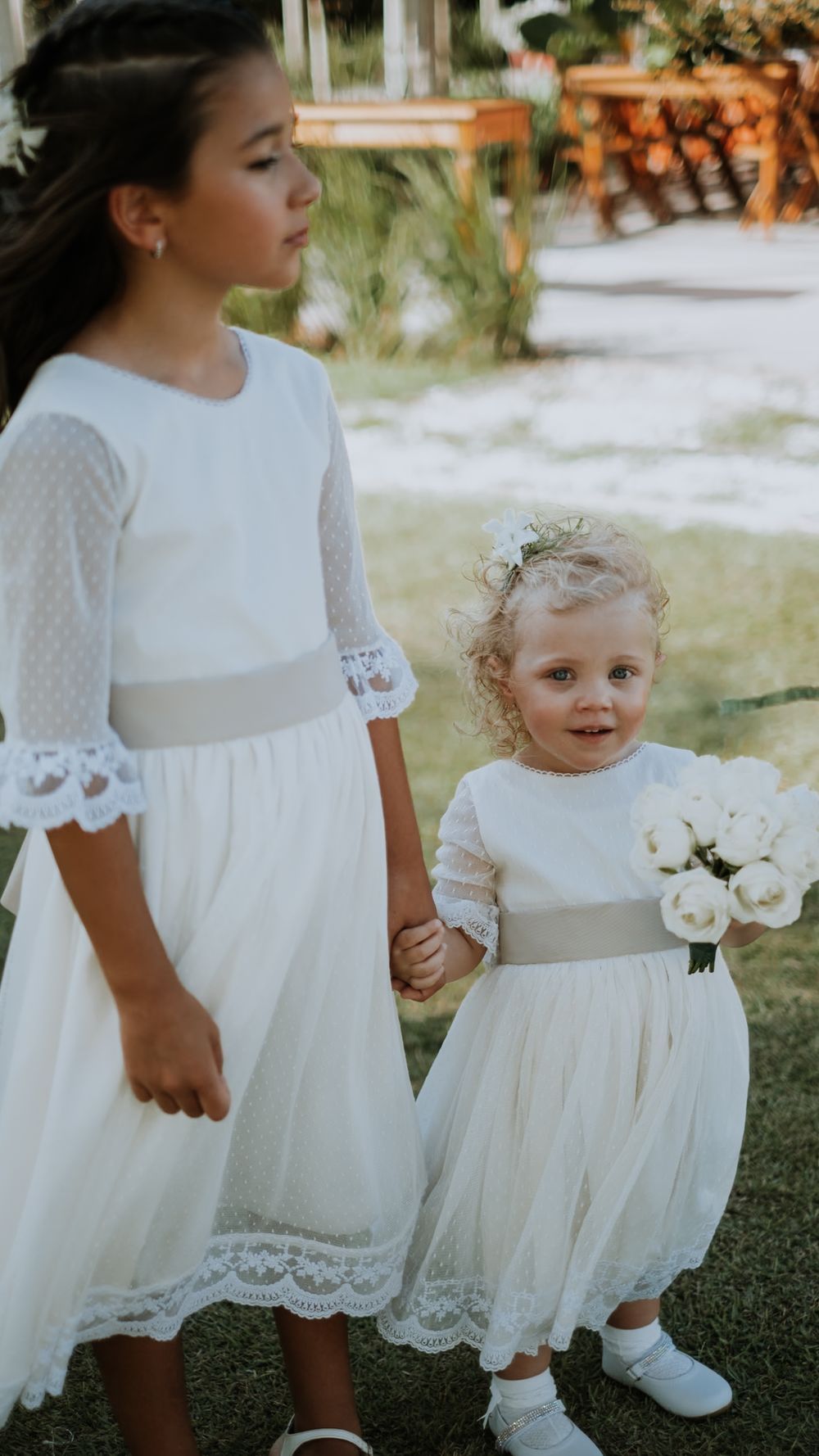Two young flower girls in white lace dresses hold hands at an outdoor wedding ceremony venue.