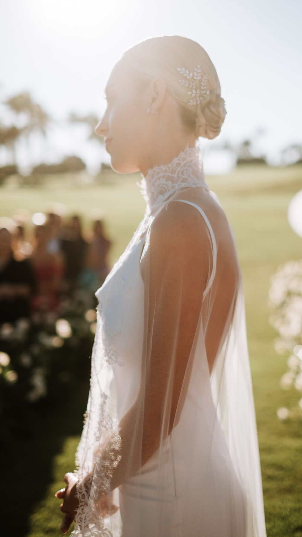 Bride in elegant backless lace high-neck wedding gown with veil and floral hair accessory at outdoor ceremony.
