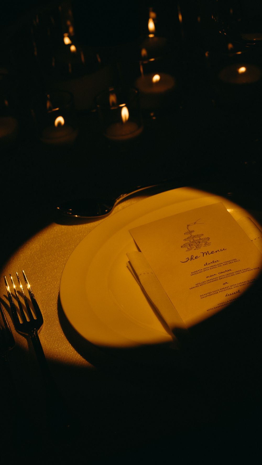 Elegant candlelit wedding place setting with white plate, fork, and menu card glowing in warm light.