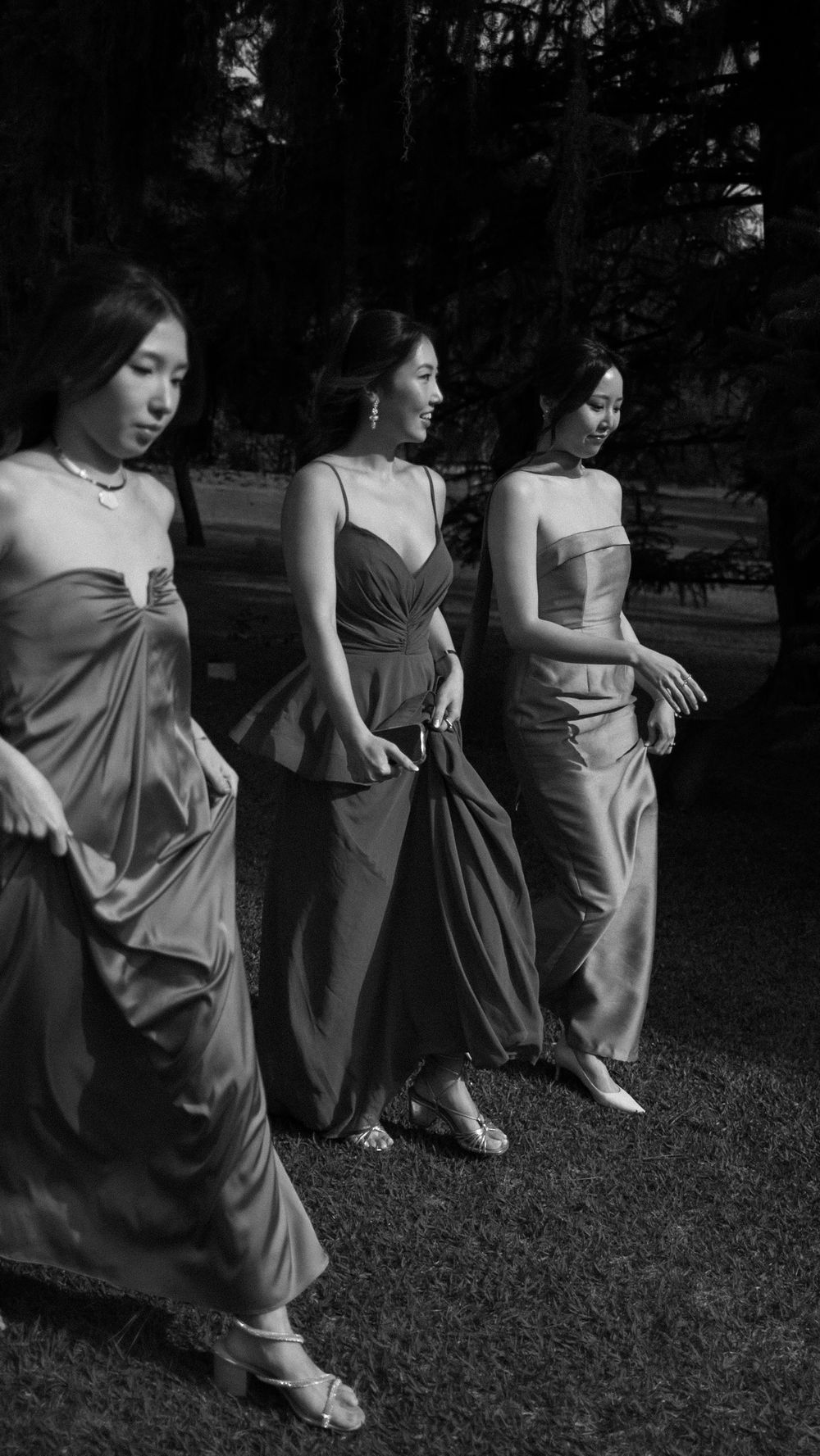 Three bridesmaids in elegant satin gowns walk gracefully outdoors in black and white wedding photography.