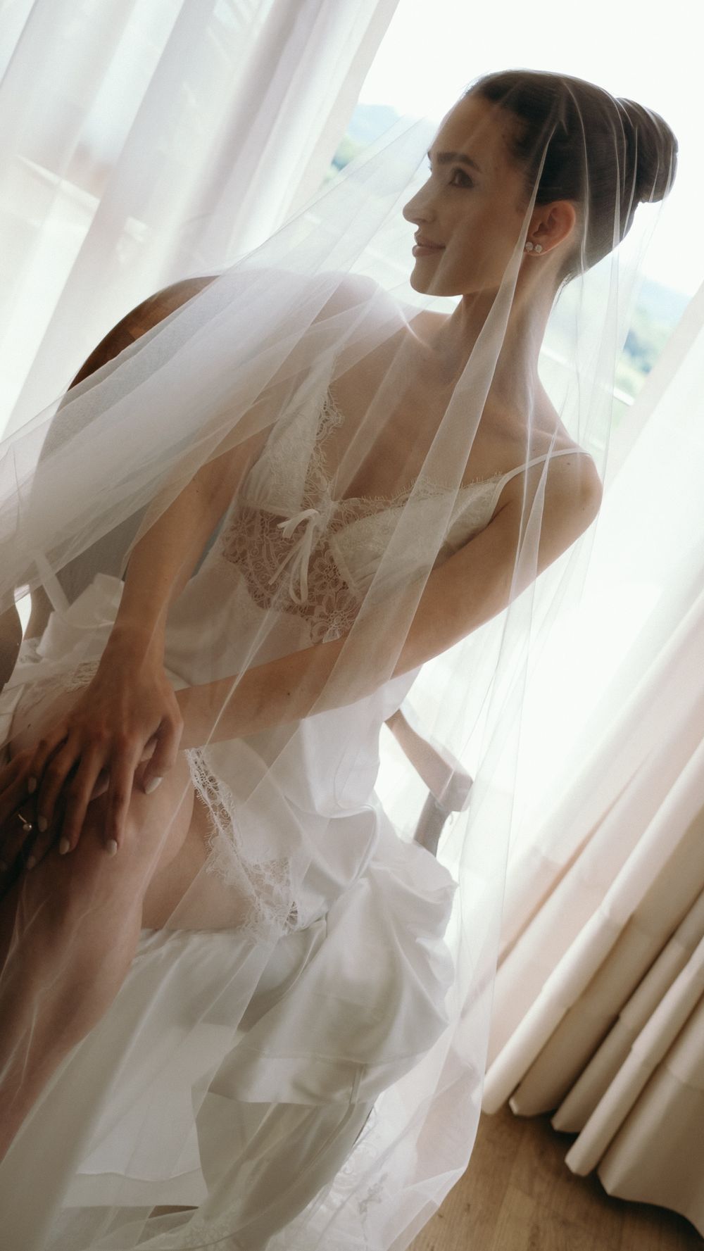 Elegant bride in white lace lingerie and veil sitting by a sunlit window before wedding.