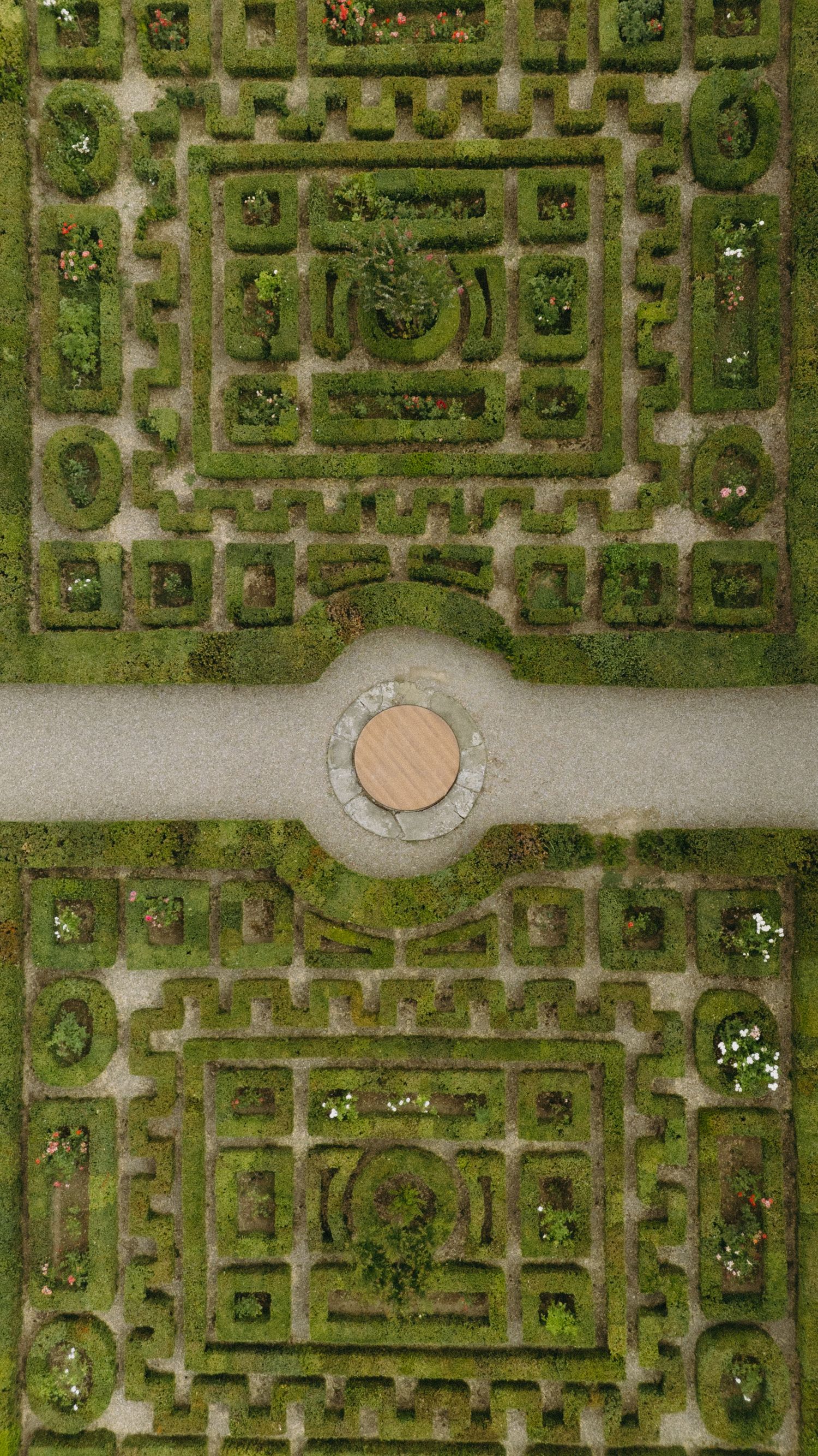 Aerial view of a symmetrical formal garden maze with intricate green hedgerows and a circular stone centerpiece.