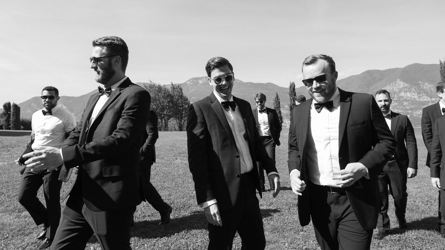Three men in formal black tuxedos walk outdoors at a wedding event with mountains in the background.