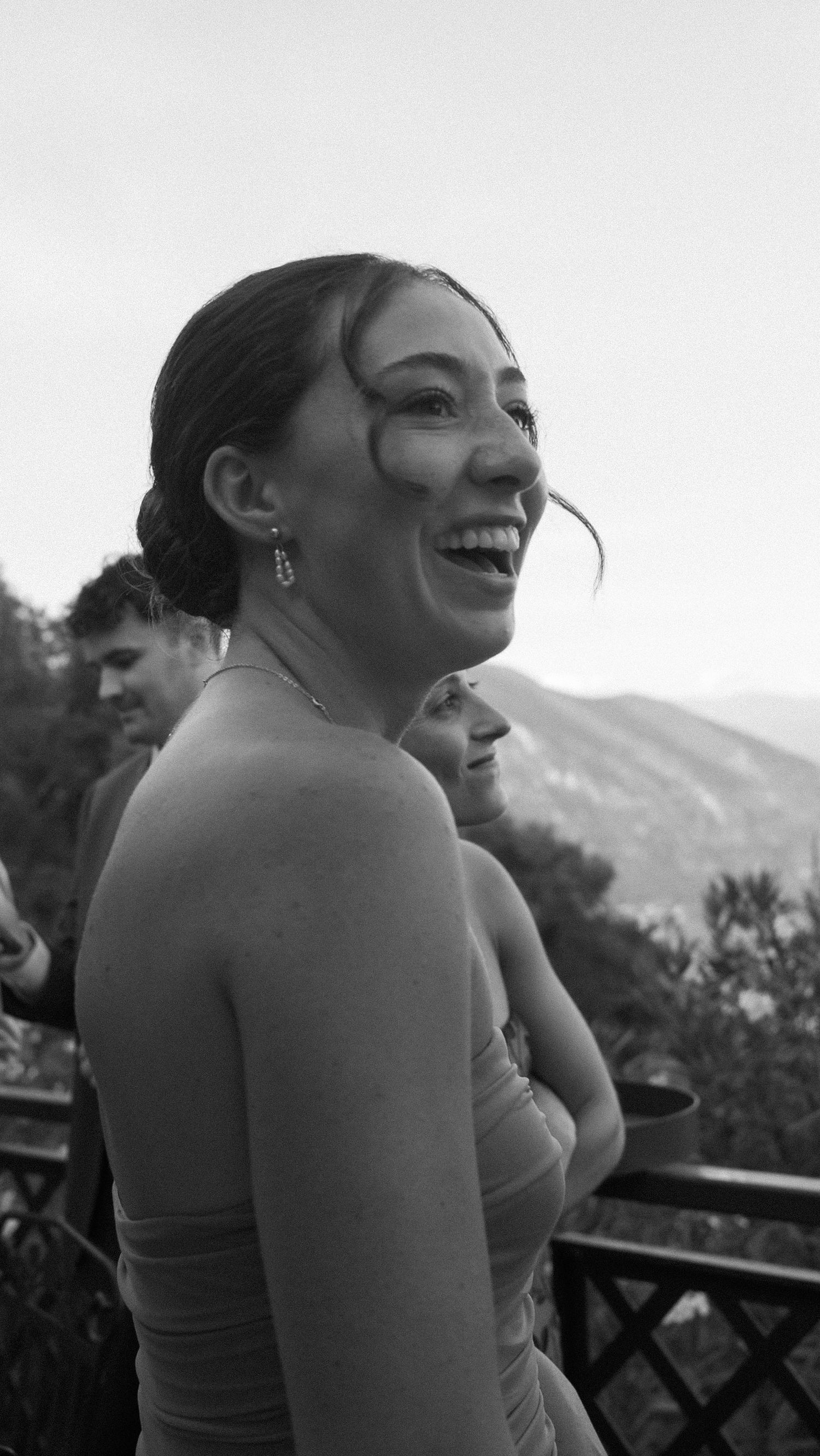 Happy young woman laughing outdoors at a wedding reception with mountain views in black and white.