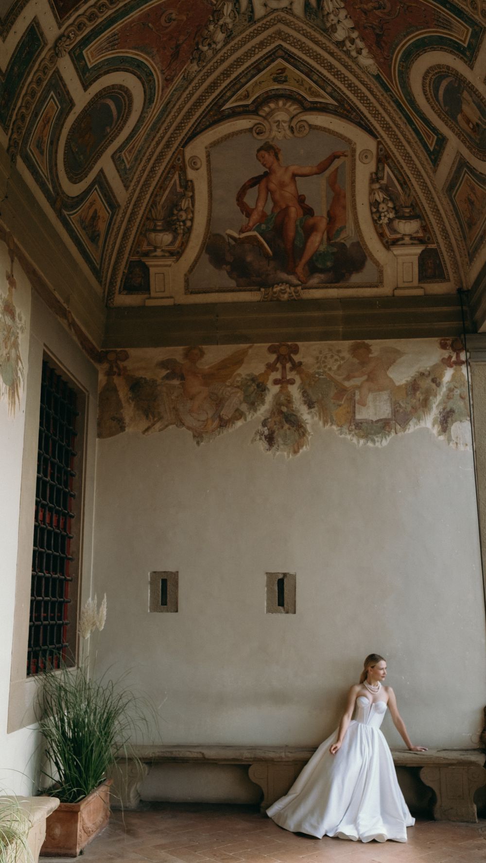 Bride in white gown stands in ornate Italian villa with Renaissance frescoes on vaulted ceiling.