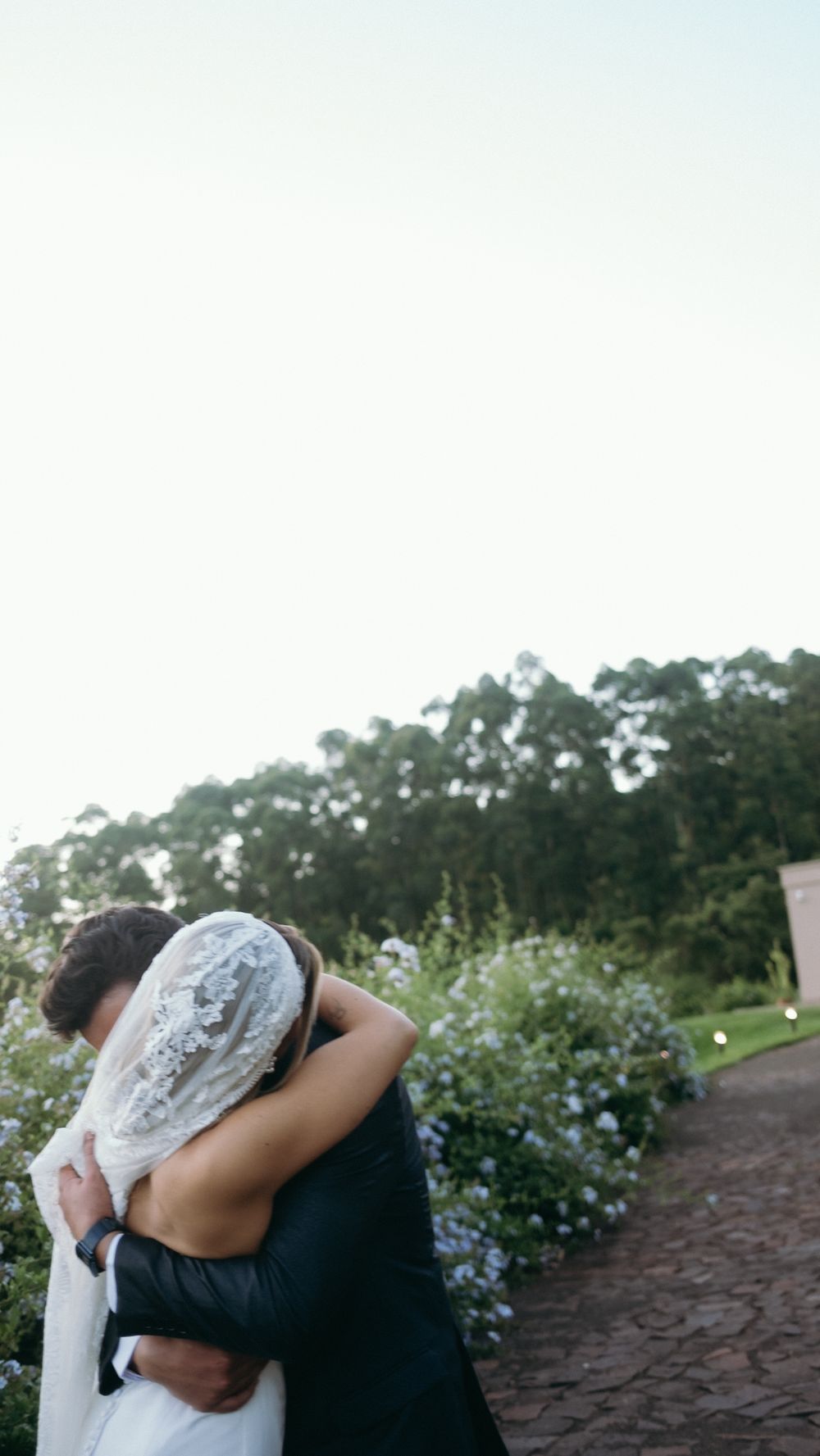 Bride and groom share a tender embrace on a garden path surrounded by blooming flowers and trees.