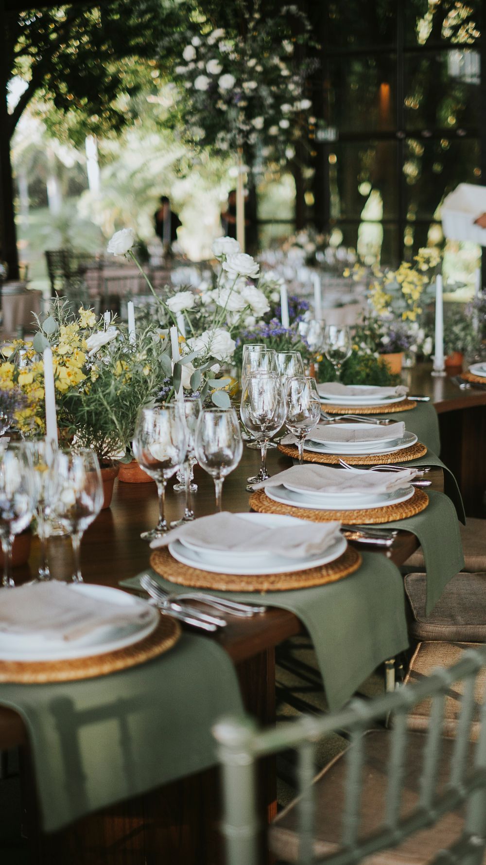 Outdoor wedding reception table with sage runner, wicker chargers, white flowers, and candles among trees.