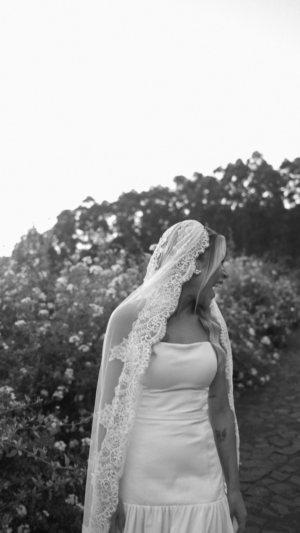 Black and white portrait of a bride wearing an intricate lace veil standing in a garden setting.