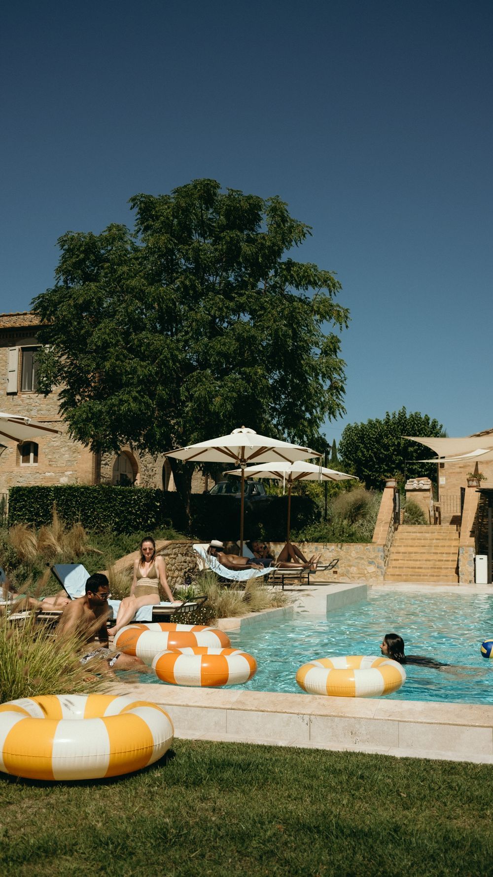 People relaxing by a sunny outdoor pool with orange and white inflatable rings near a rustic stone villa.