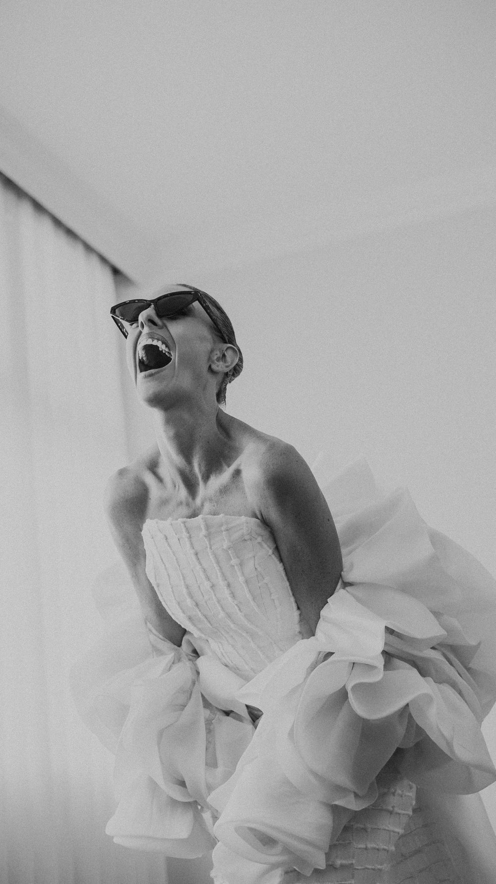 Joyful bride in ruffled white strapless gown laughs while wearing sunglasses in black and white photo.