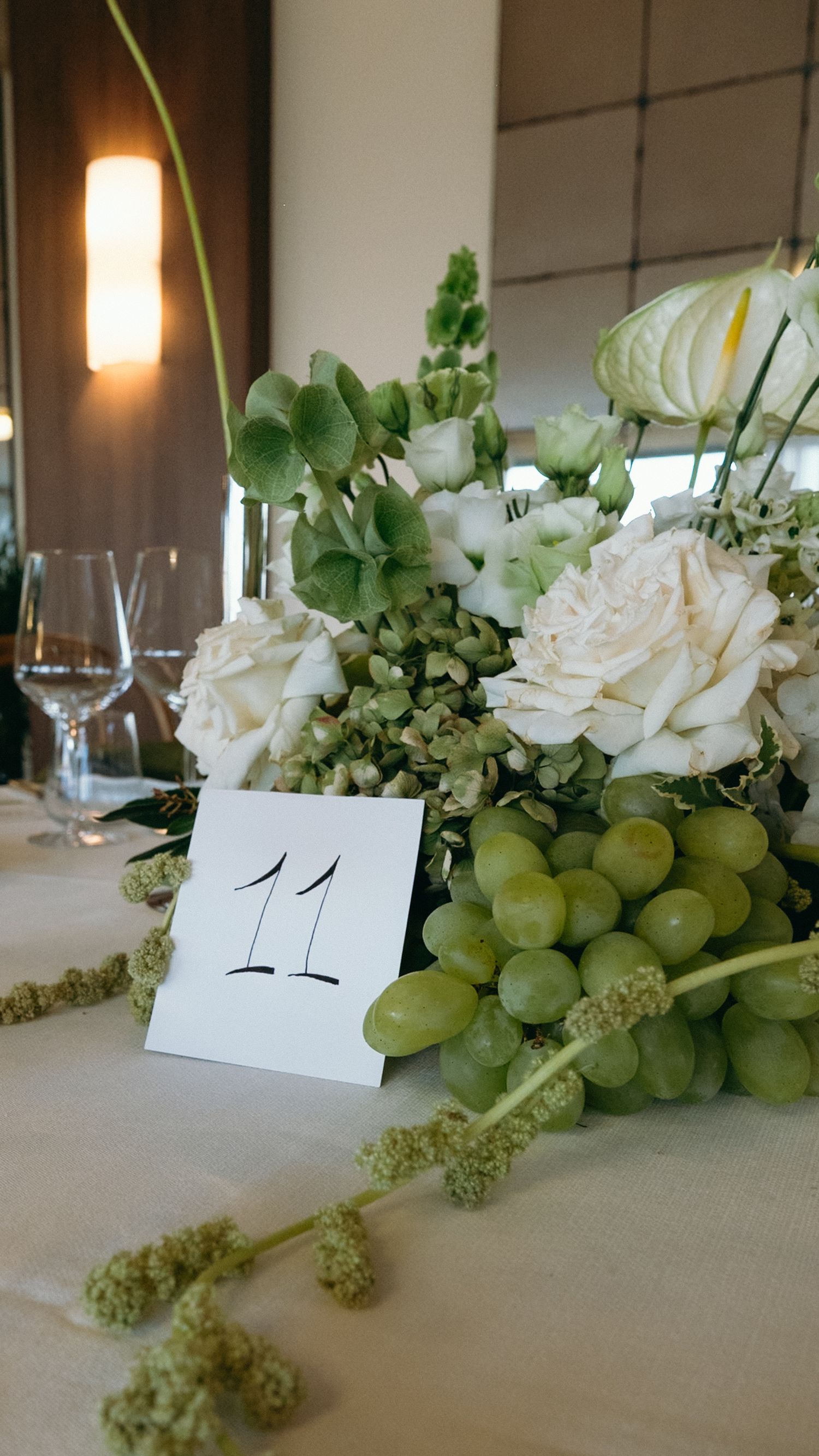 Elegant wedding table centerpiece with white roses, green hydrangeas, grapes, and a number 11 card.