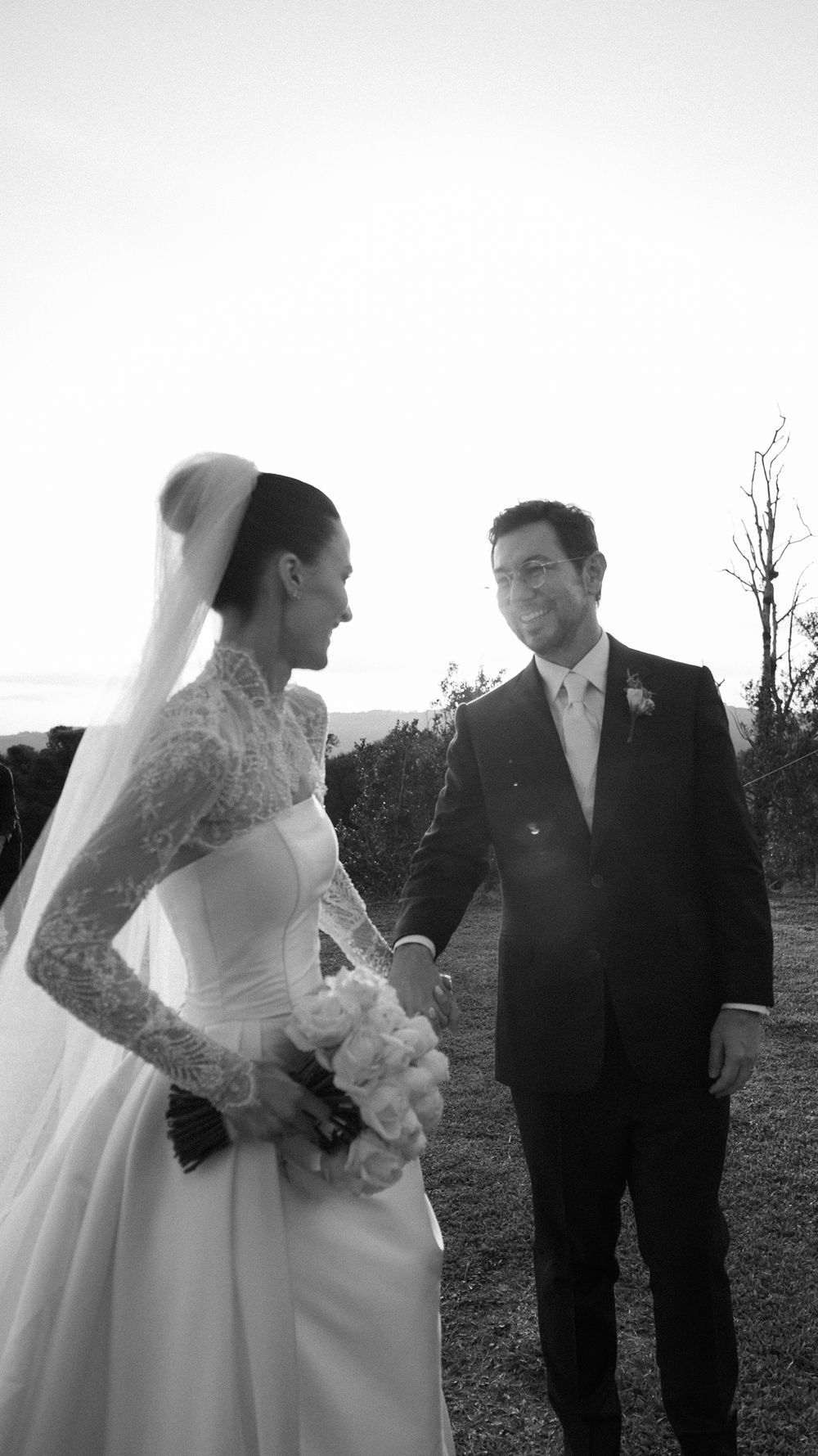 Bride and groom hold hands outdoors at sunset in elegant black and white wedding portrait.