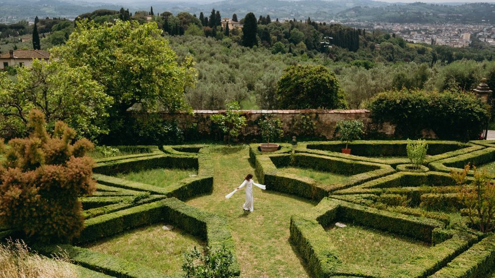 Bride in white wedding dress walking through symmetrical Italian garden maze on a sunny day.