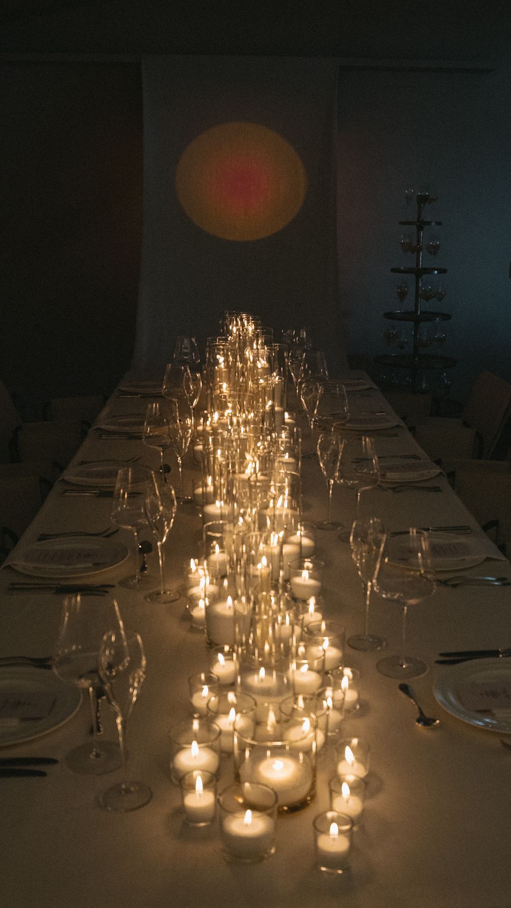 Long wedding reception table illuminated by rows of twinkling candles with glassware and circular light projection.