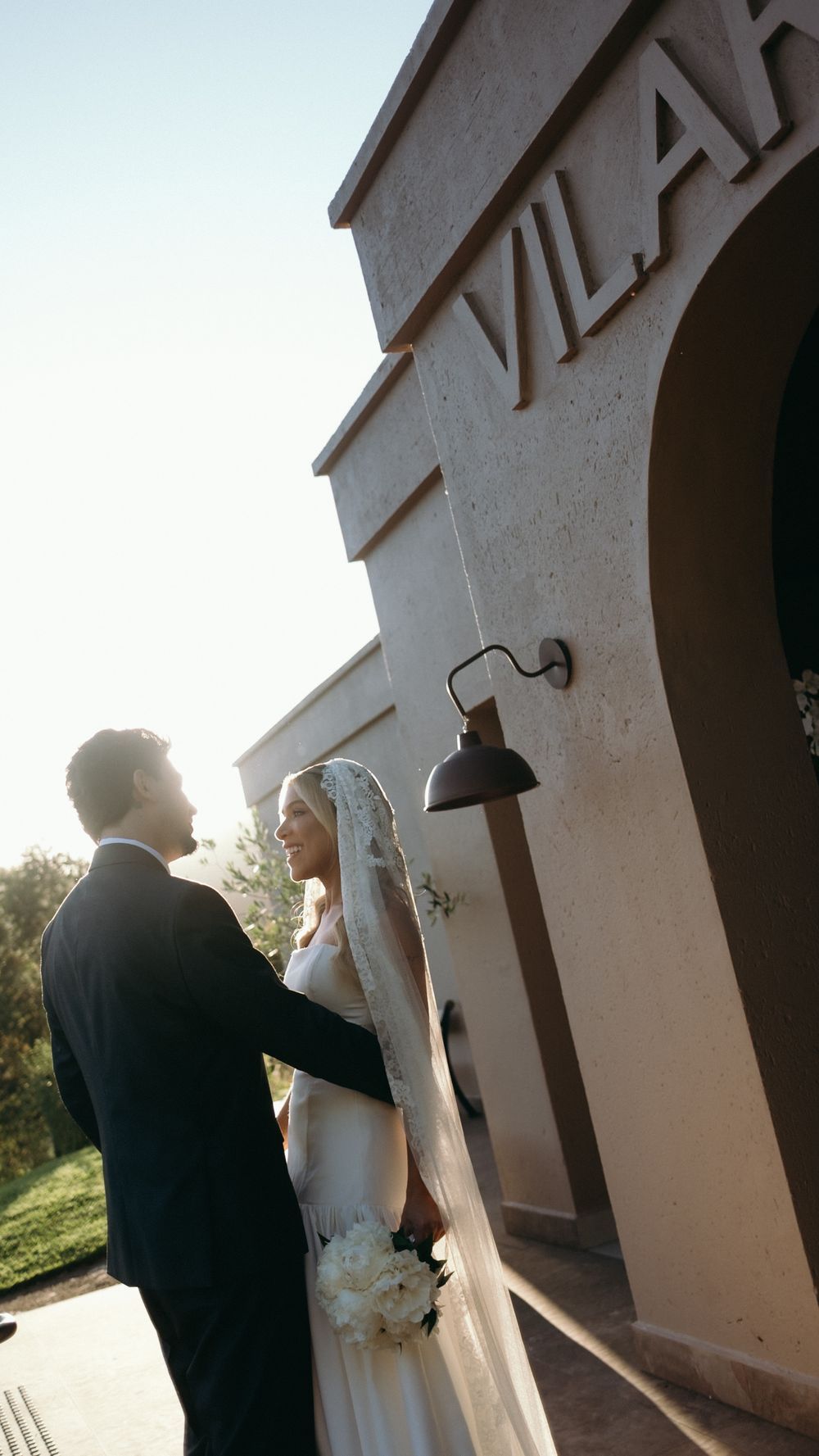 Bride and groom share a romantic moment outside a venue with VILAH signage in warm golden sunlight.