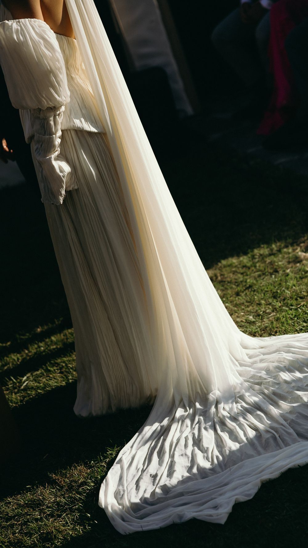 Close-up of bride in flowing white pleated wedding gown with long train and elegant gloves on grass.