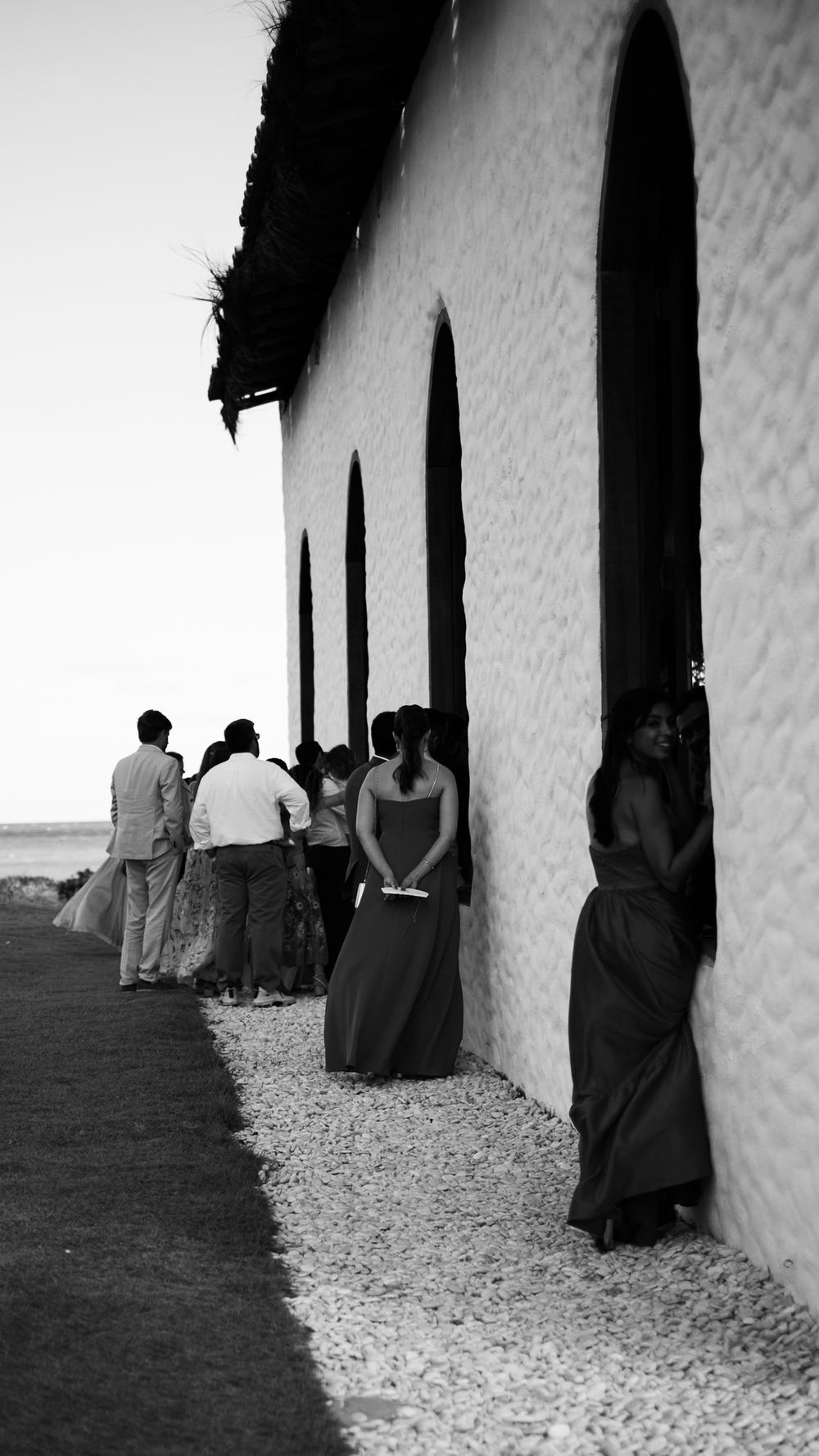 Black and white photo of wedding guests and bridesmaids gathered outside a whitewashed chapel with arched windows.