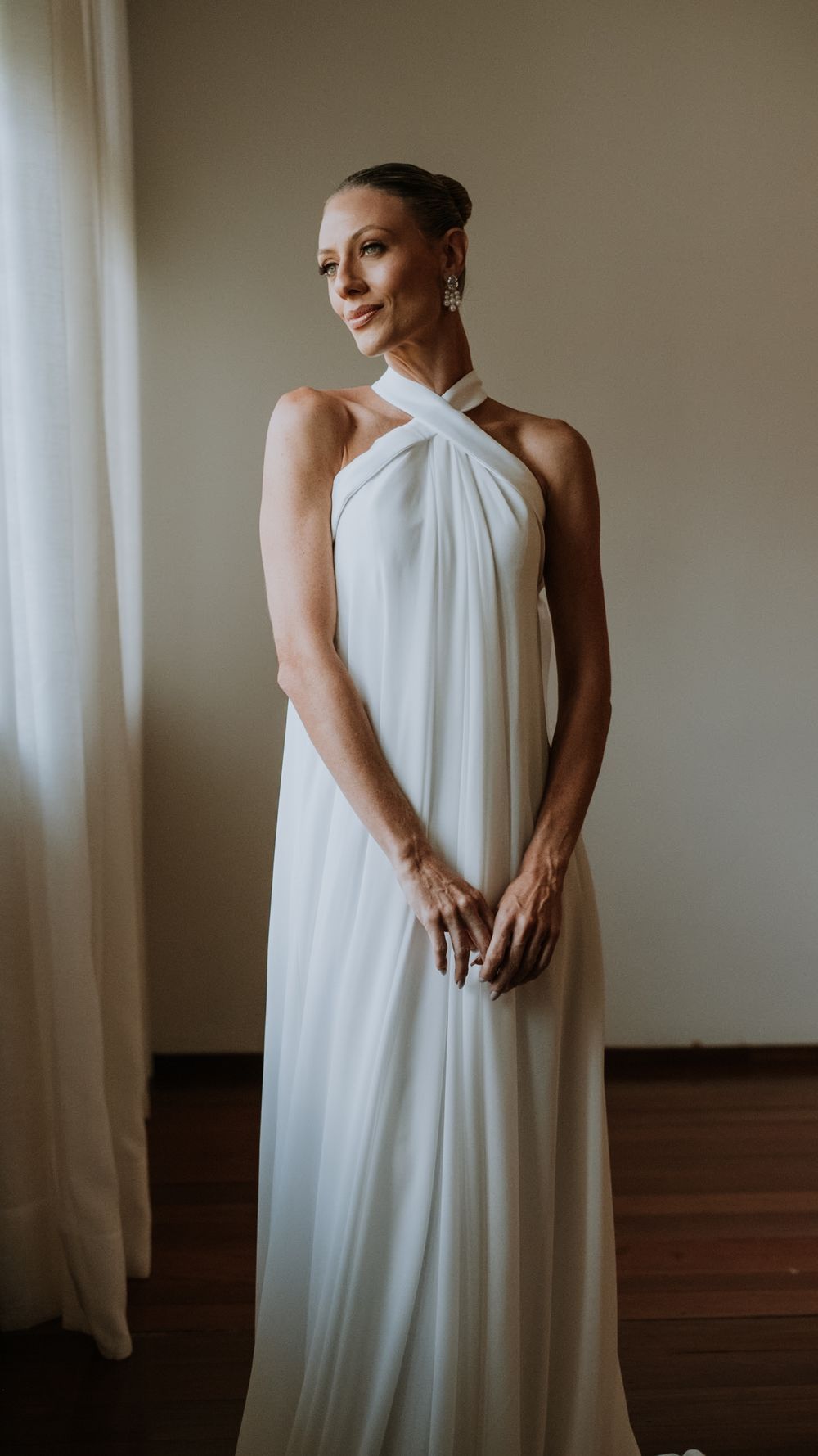 Elegant bride in minimalist white halter-neck draped wedding gown poses near window in soft light.