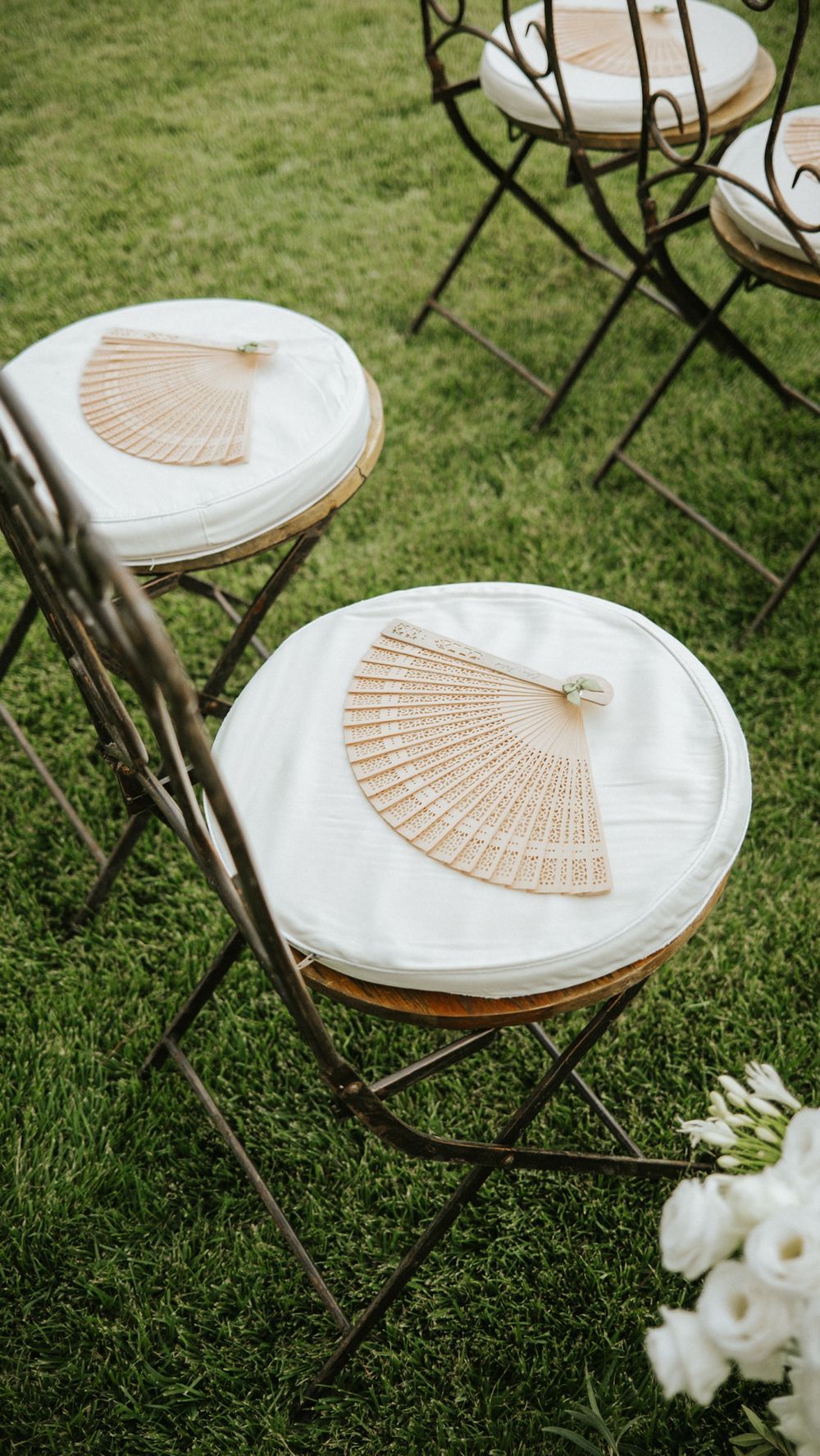 Wooden hand fans placed on white cushioned chairs arranged on grass for outdoor wedding ceremony.