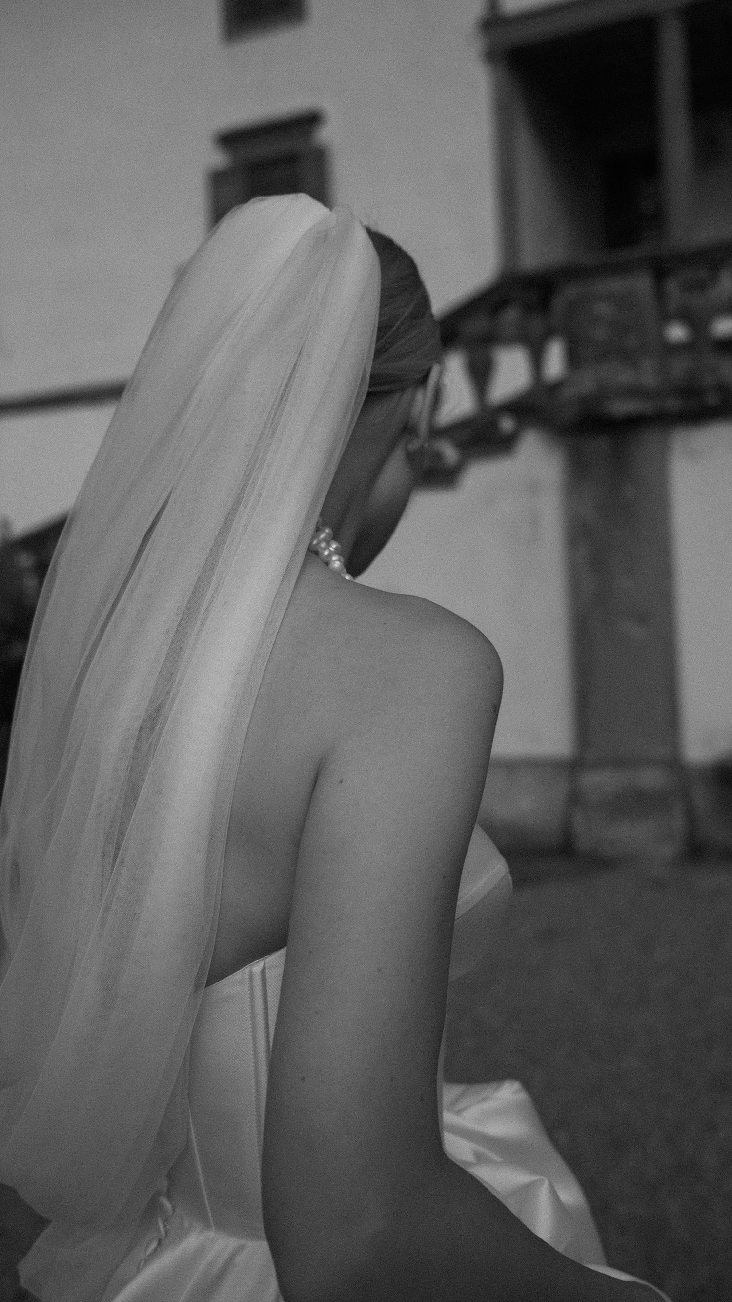 Black and white photo of a bride in a backless wedding dress and veil, viewed from behind outdoors.