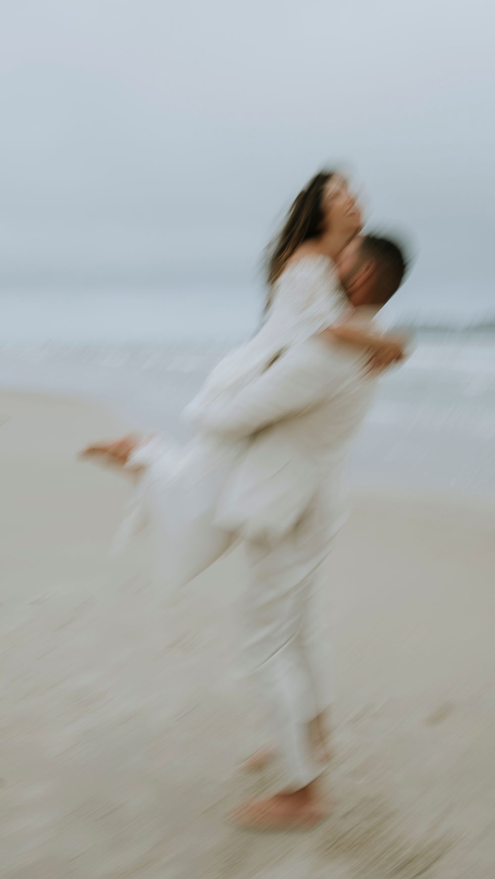 Blurred motion shot of a couple embracing on a beach in matching white outfits.