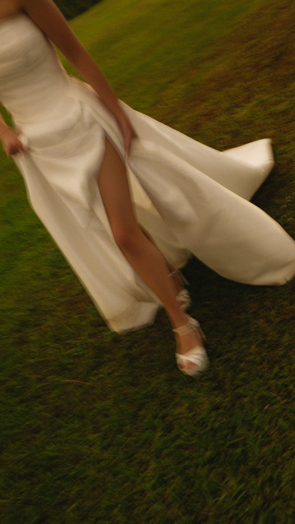 Close-up of bride lifting white wedding gown while walking across lush green grass in white heeled sandals.
