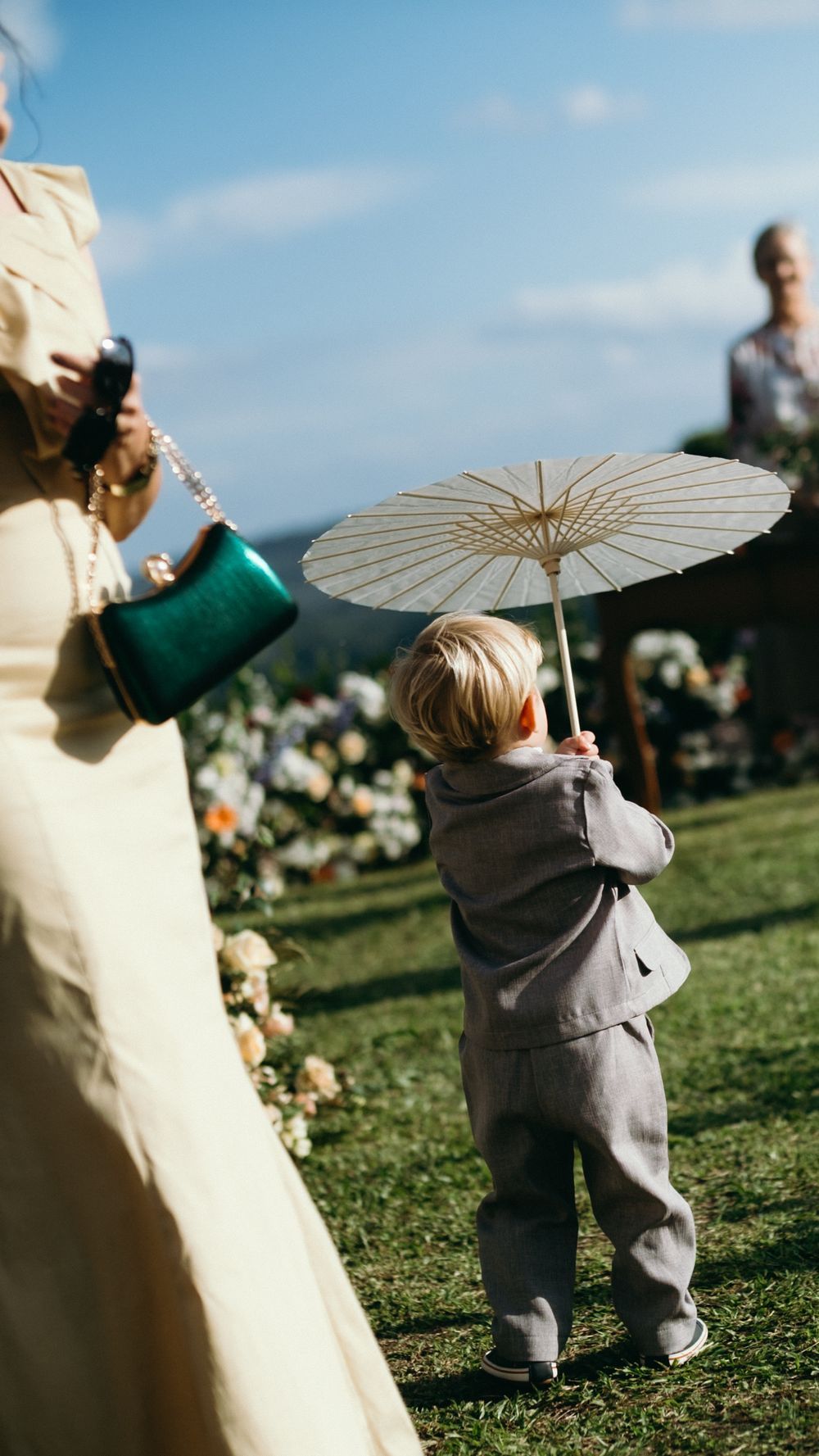 Adorable young child in grey suit holding a white parasol umbrella at an outdoor wedding ceremony.